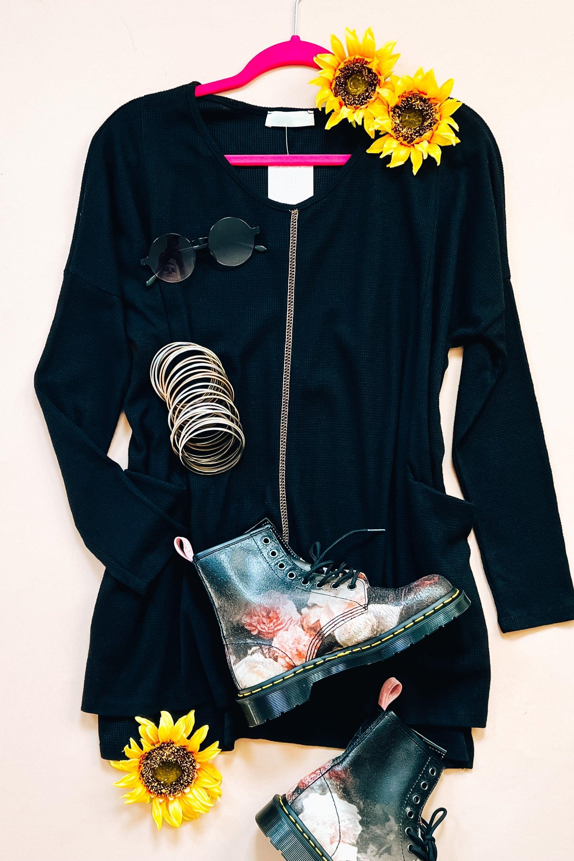 Black long-sleeve tunic with zipper detail styled with floral print boots, round sunglasses, bangles, and sunflowers on a pale background