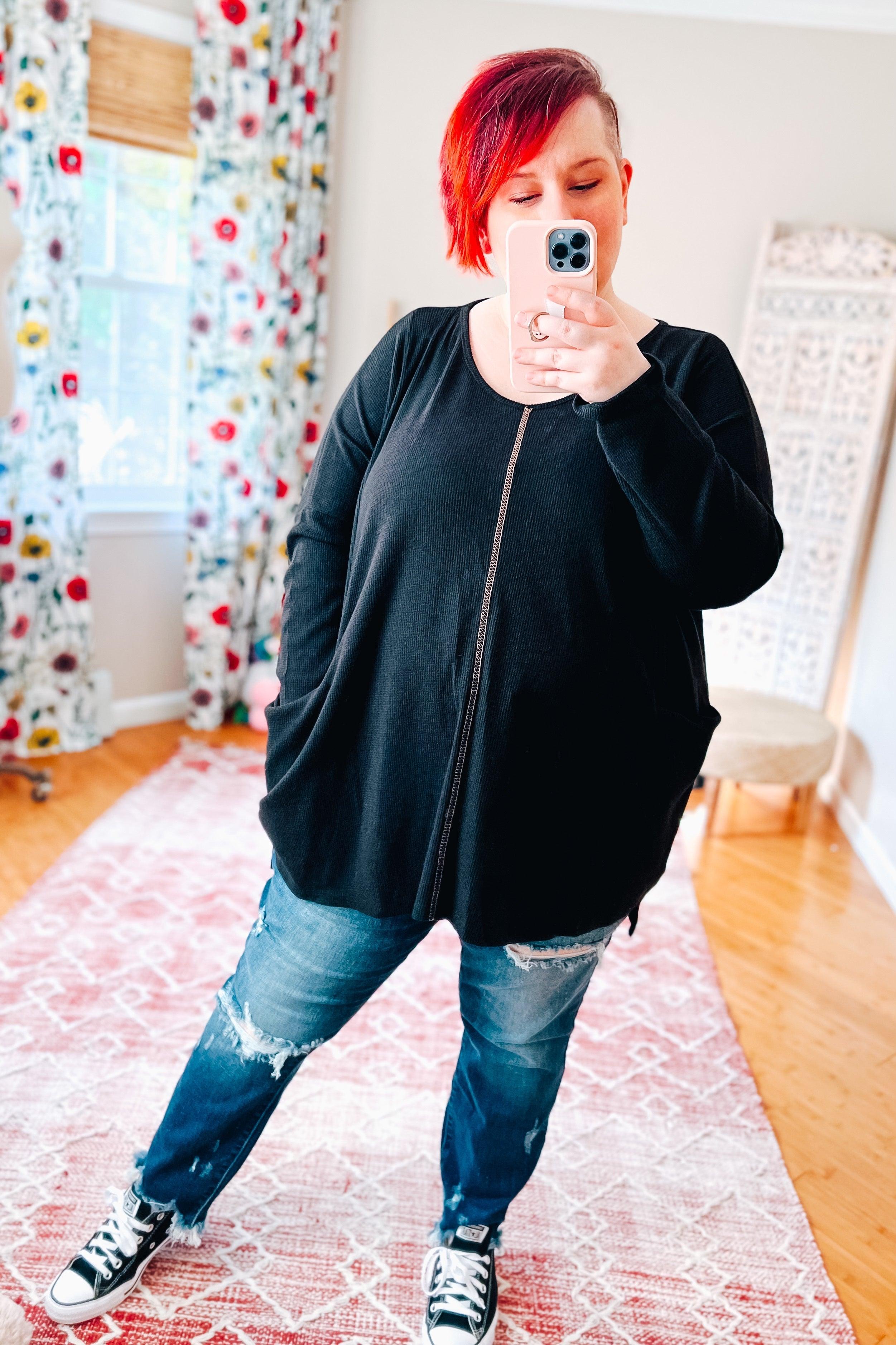 Woman taking a selfie wearing a loose black thermal tunic with pockets and ripped blue jeans in a bright room