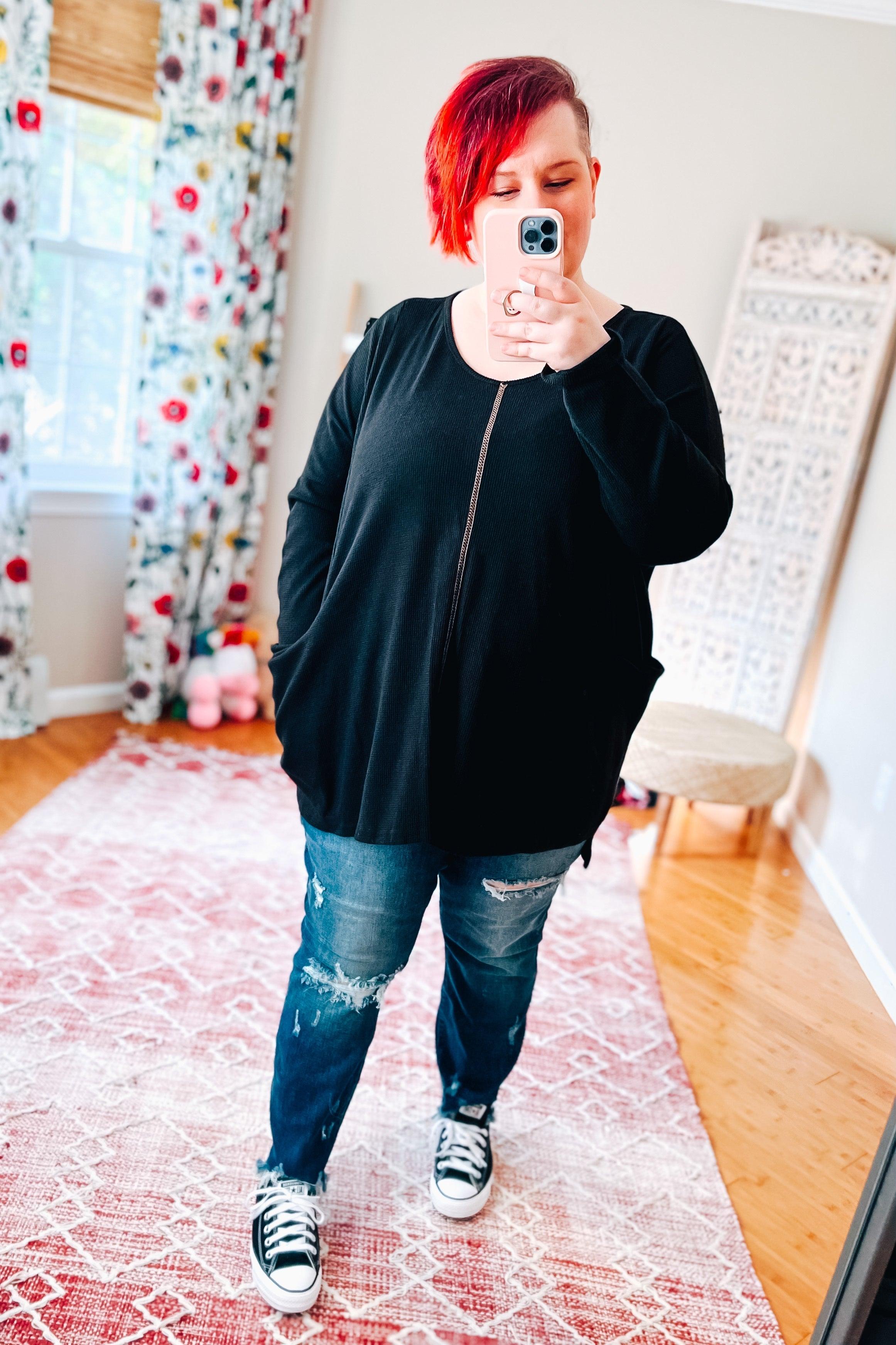 Woman taking mirror selfie wearing a loose black long-sleeve tunic and ripped blue jeans in a bright room