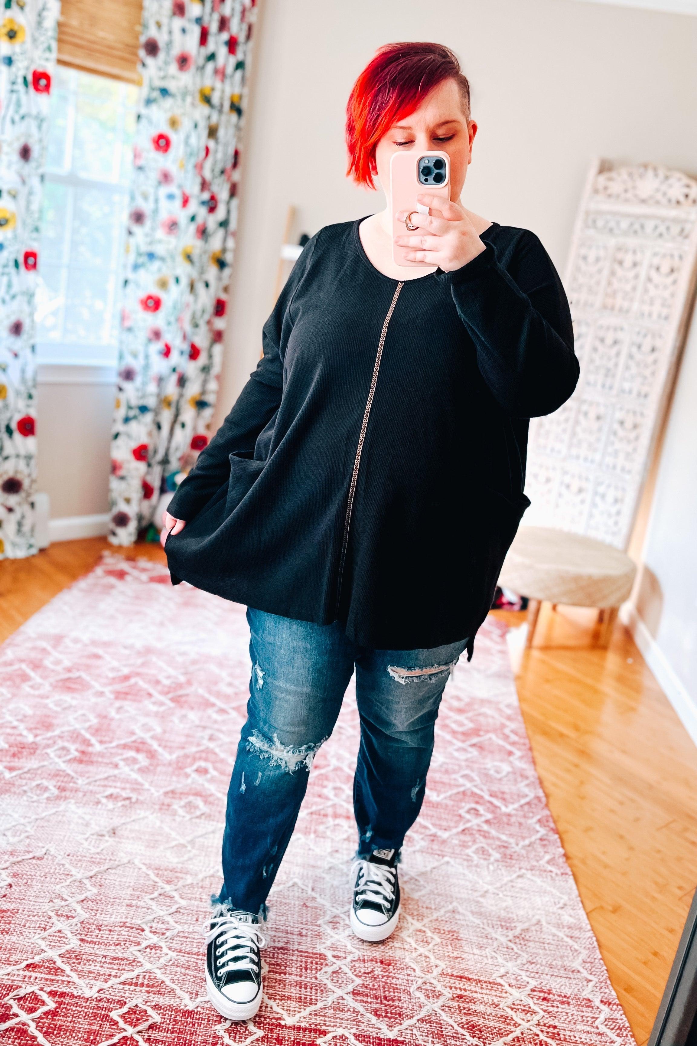 Woman wearing a loose black tunic with front seam detail and distressed blue jeans standing indoors on a patterned rug