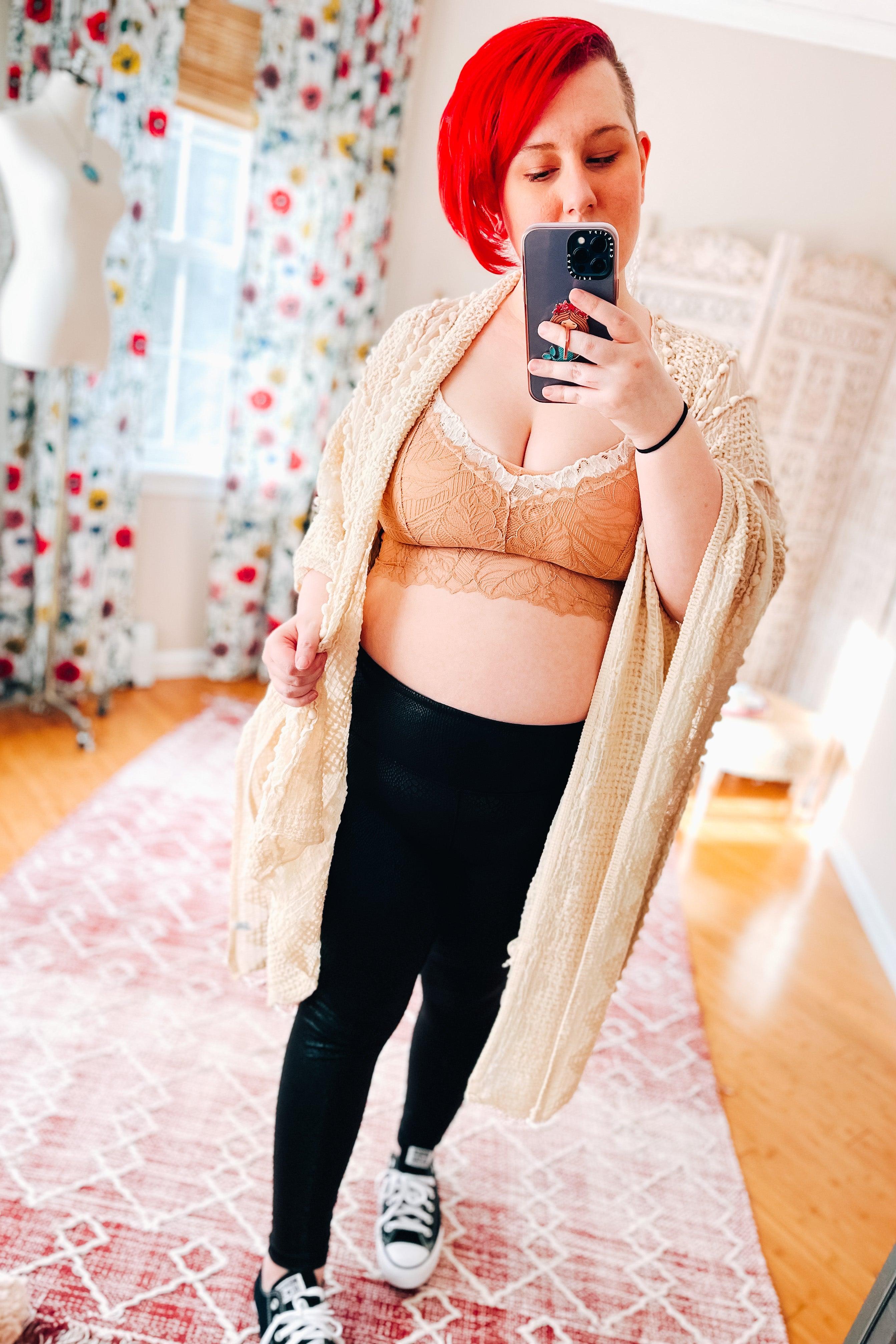 Woman with bright red hair wearing a brick-colored lace bralette with scalloped edges, black leggings, and a cream knitted cardigan, taking a mirror selfie in a sunlit room.