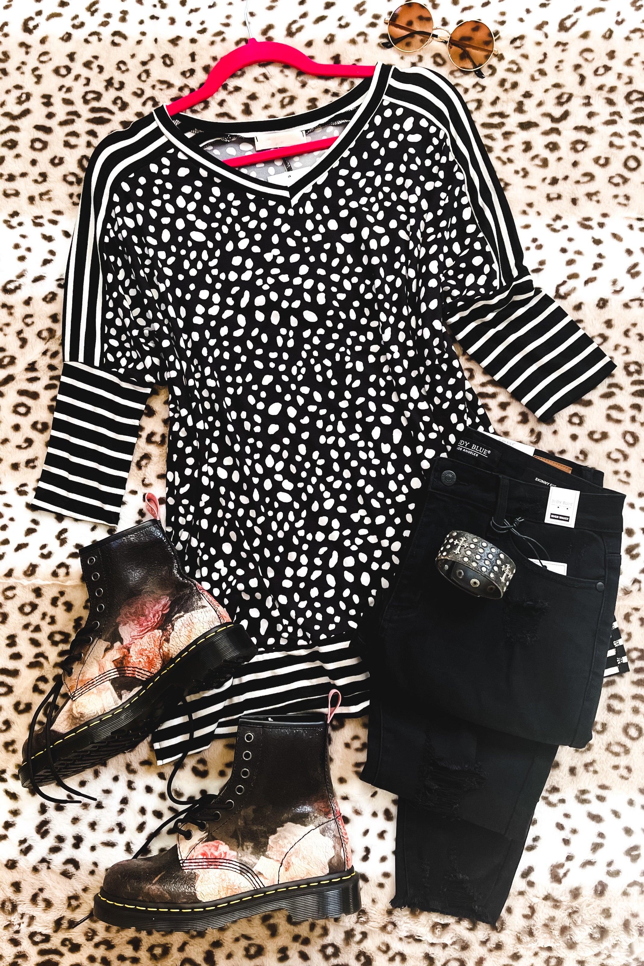Black tunic with white irregular spots and striped sleeves laid out with black distressed jeans, studded belt, floral lace-up boots, and round tortoiseshell sunglasses on leopard print fabric background.