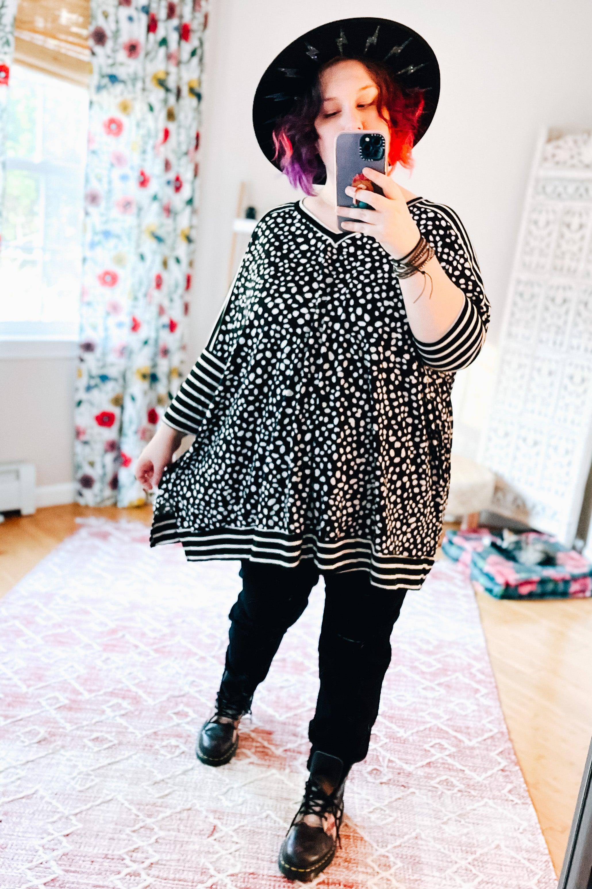Woman wearing a loose black and white polka dot tunic with striped mid sleeves and hem, black pants, black boots, and a wide-brim black hat taking a mirror selfie in a bright room.