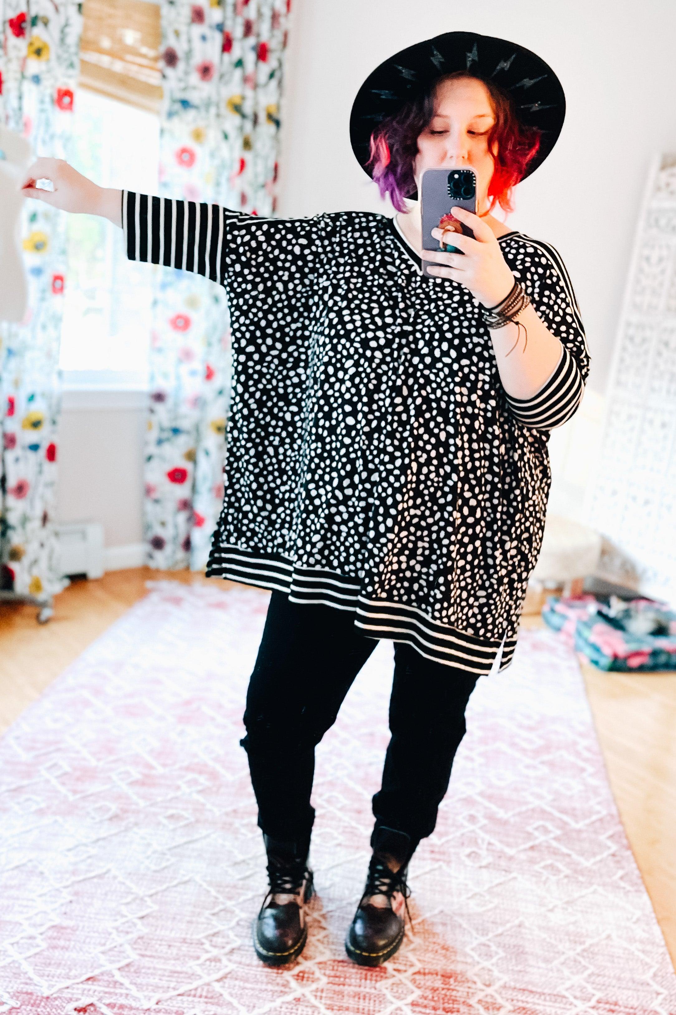 Woman wearing a loose black tunic with white abstract spots and contrasting striped sleeves and hem, black pants, black boots, and a black hat taking a mirror selfie in a bright room.