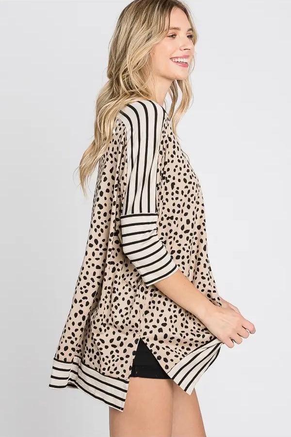 Side view of a woman smiling, wearing a taupe tunic featuring black irregular spots and contrasting black and white horizontal stripes on the sleeves and hem