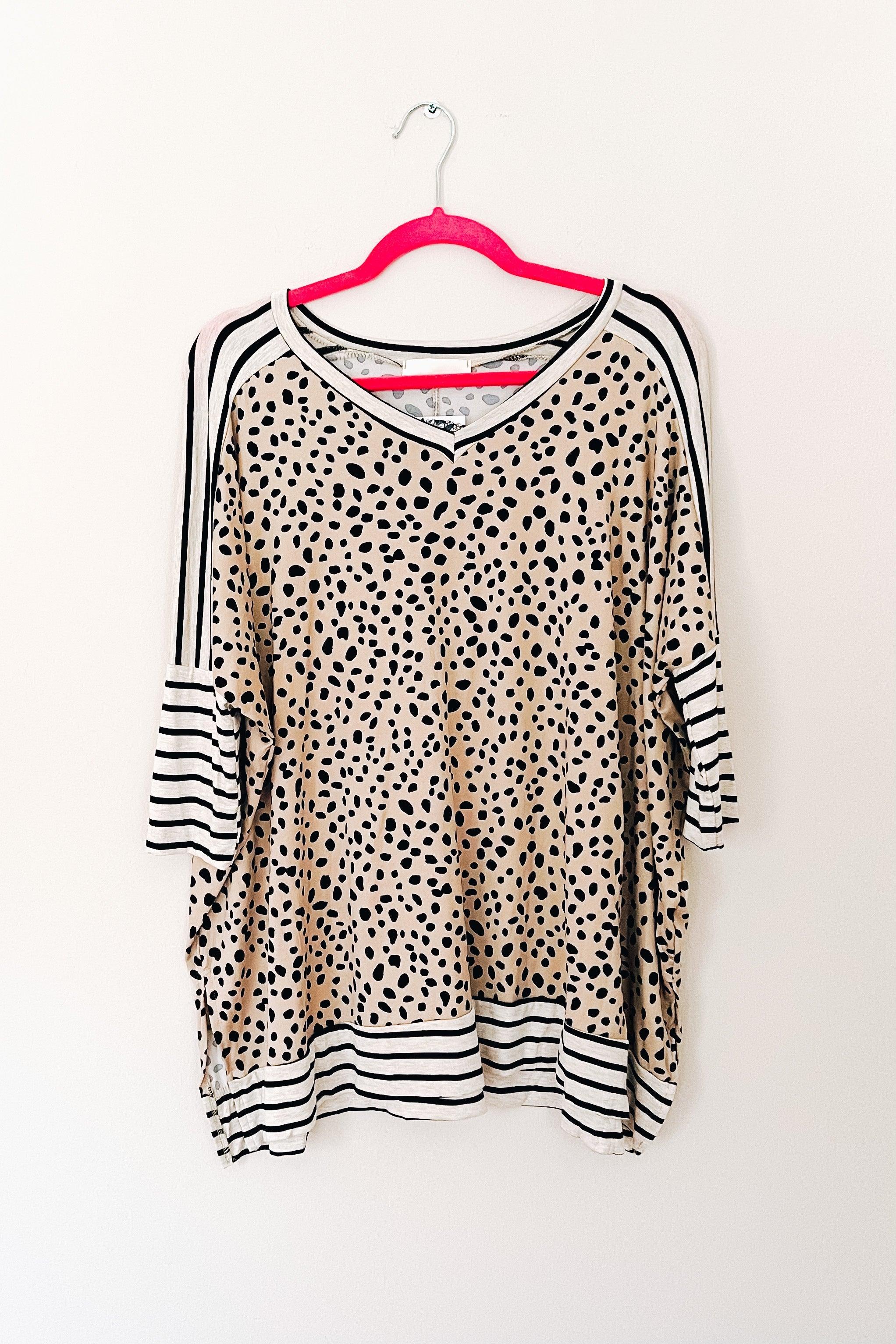 Loose taupe tunic with scattered black spots and contrasting black and white horizontal stripes on sleeves, neckline, and hem, hanging on a hanger.