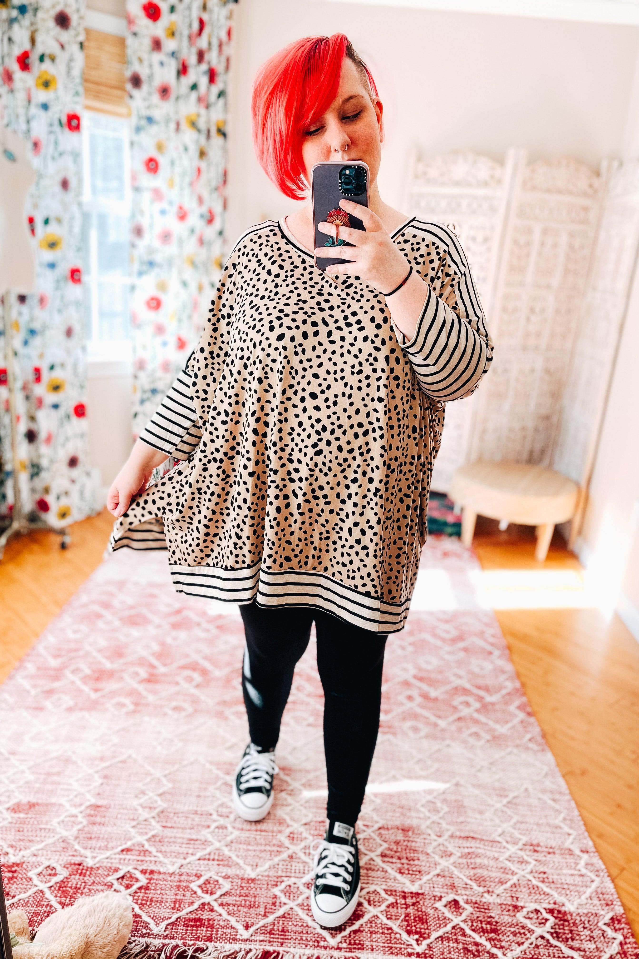Woman taking a mirror selfie wearing a taupe mid sleeve tunic with black irregular spots and contrasting black and white striped sleeves and hem, paired with black leggings and black sneakers.
