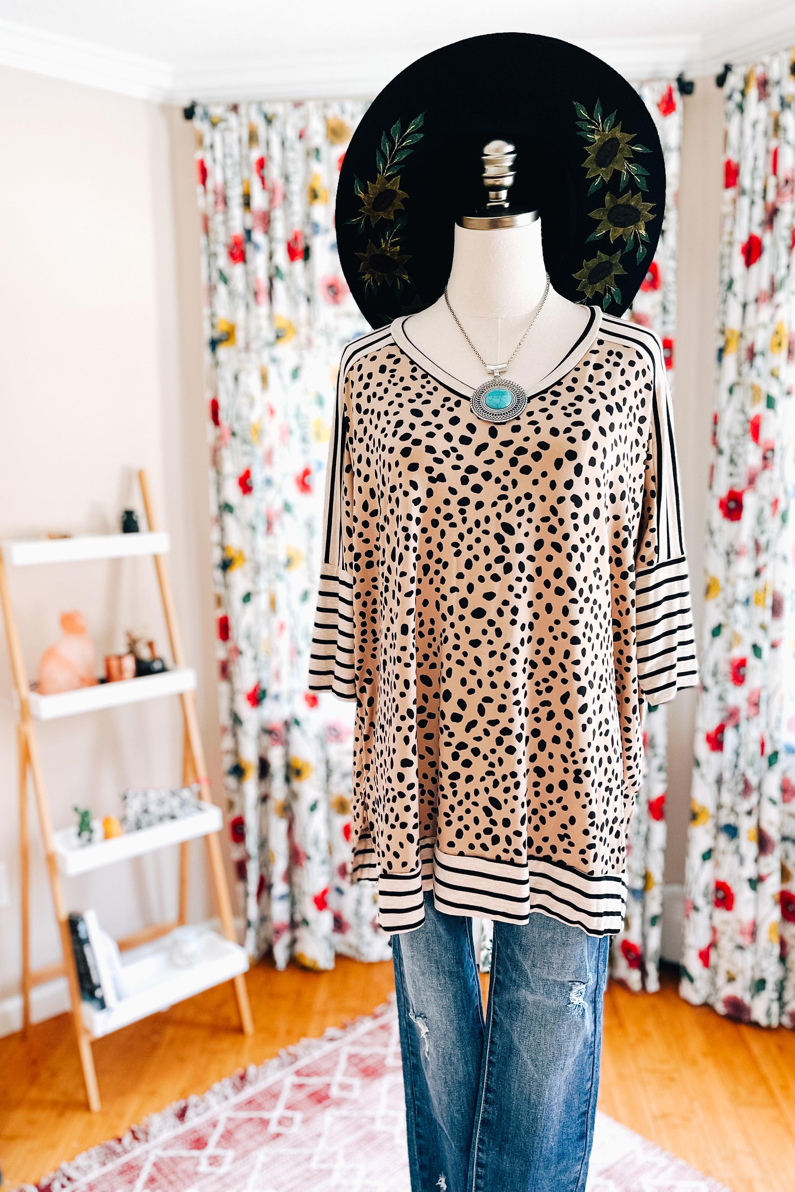 Mannequin dressed in a taupe tunic with black irregular spots and contrasting black and white stripes on the sleeves and hem, paired with distressed blue jeans and layered necklaces.