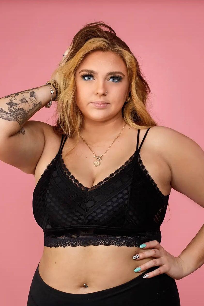 Curvy woman wearing a black lace bralette with geometric mesh and scalloped trim, posing confidently with hand in hair against a pink background.