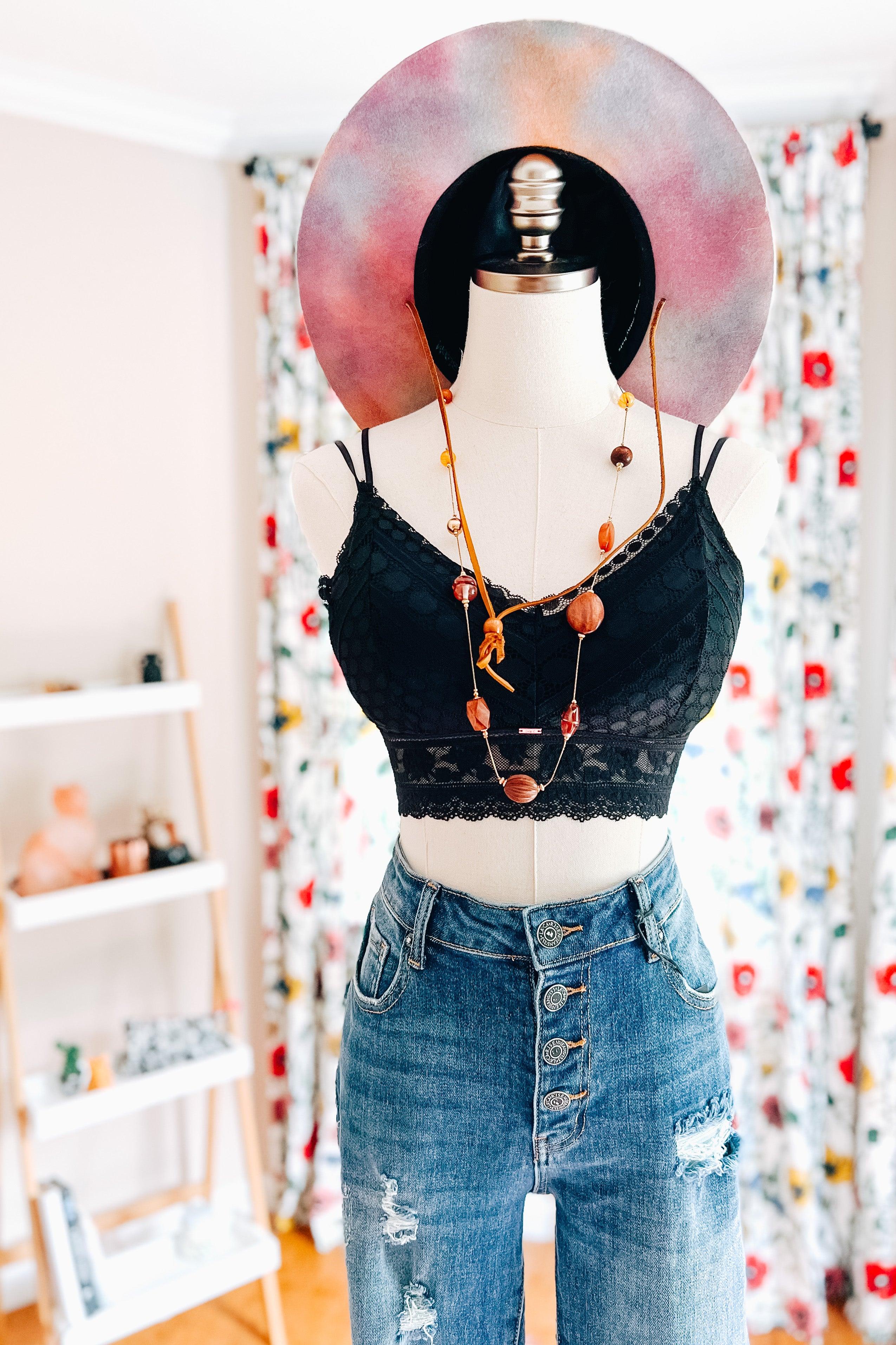Mannequin dressed in a black lace bralette with a textured pattern and scalloped trim, paired with high-waisted distressed blue jeans and layered bead necklaces, set against a bright room with floral curtains and a pastel wide-brim hat.