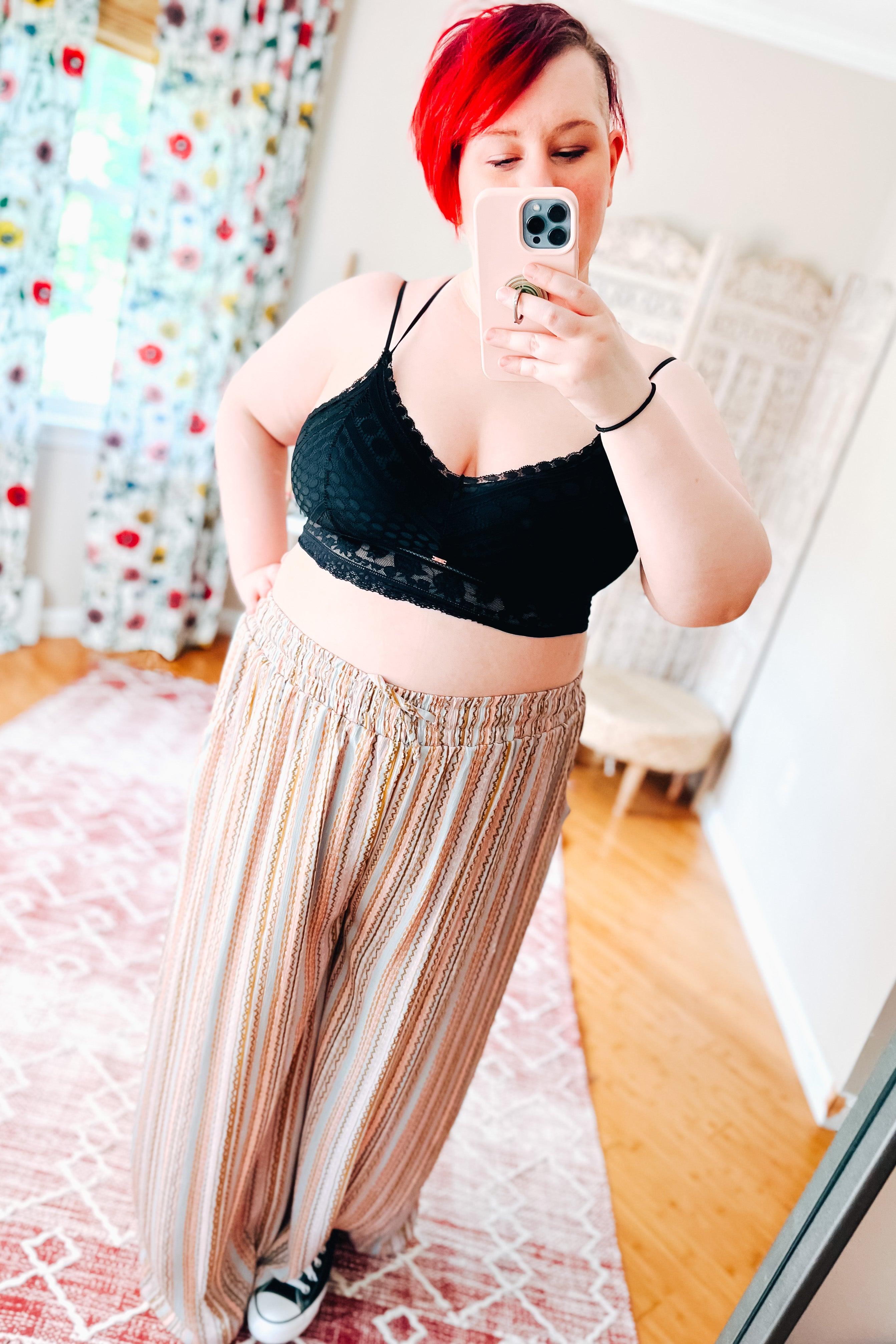 Woman with short red hair wearing a black bralette with geometric lace patterns and scalloped edges, paired with wide-leg striped pants and black sneakers, taking a mirror selfie in a bright room with floral curtains and wooden flooring.