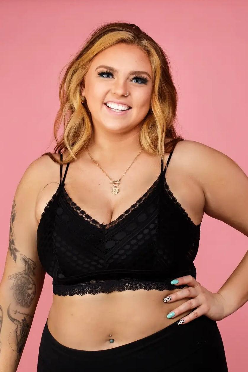 Smiling woman wearing a black lace bralette with delicate double straps and scalloped edges, posing confidently against a pink background.
