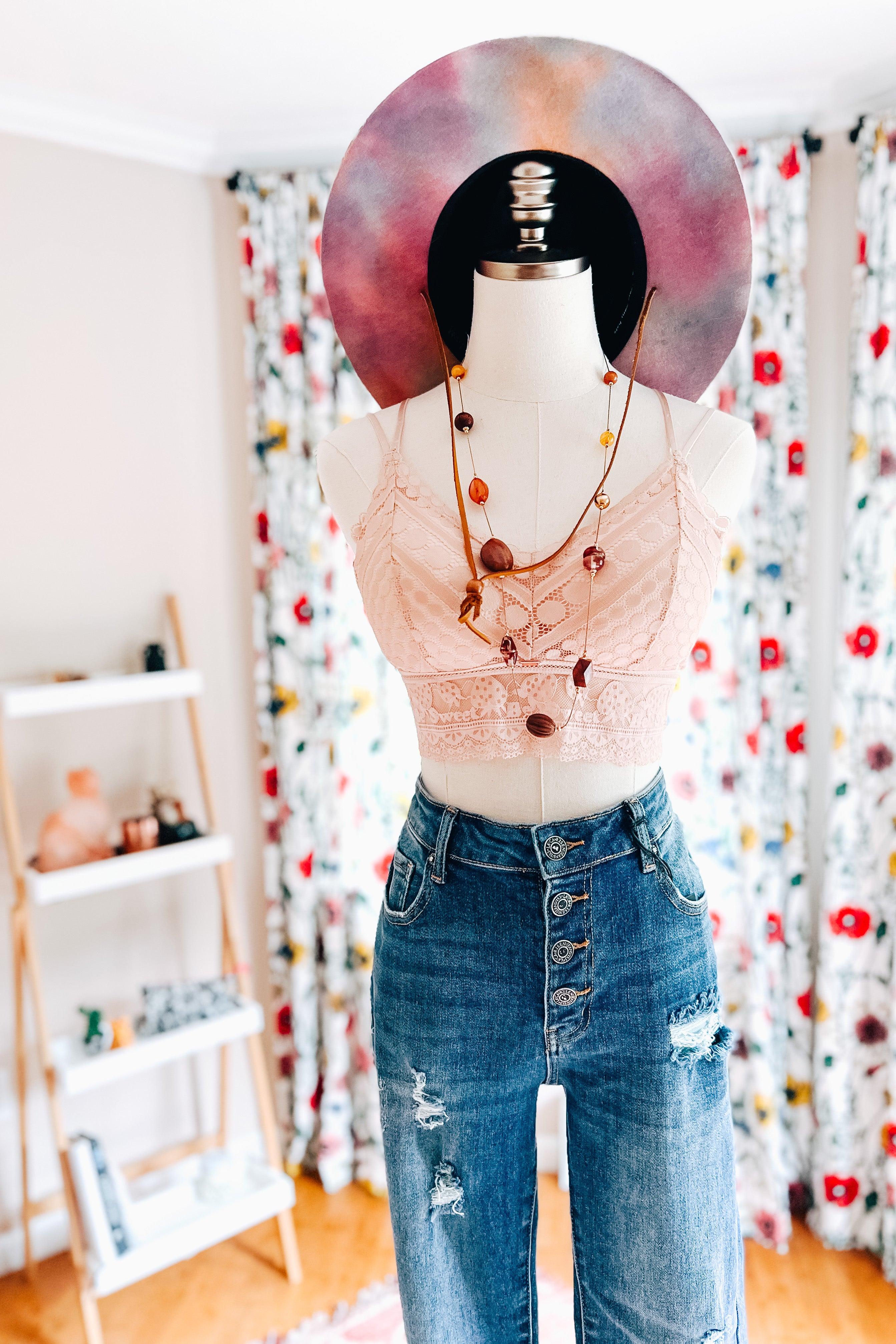 Mannequin dressed in a blush lace bralette with intricate detailing paired with high-waisted distressed denim jeans and a colorful wide-brimmed hat, styled with a beaded necklace against a floral curtain backdrop.