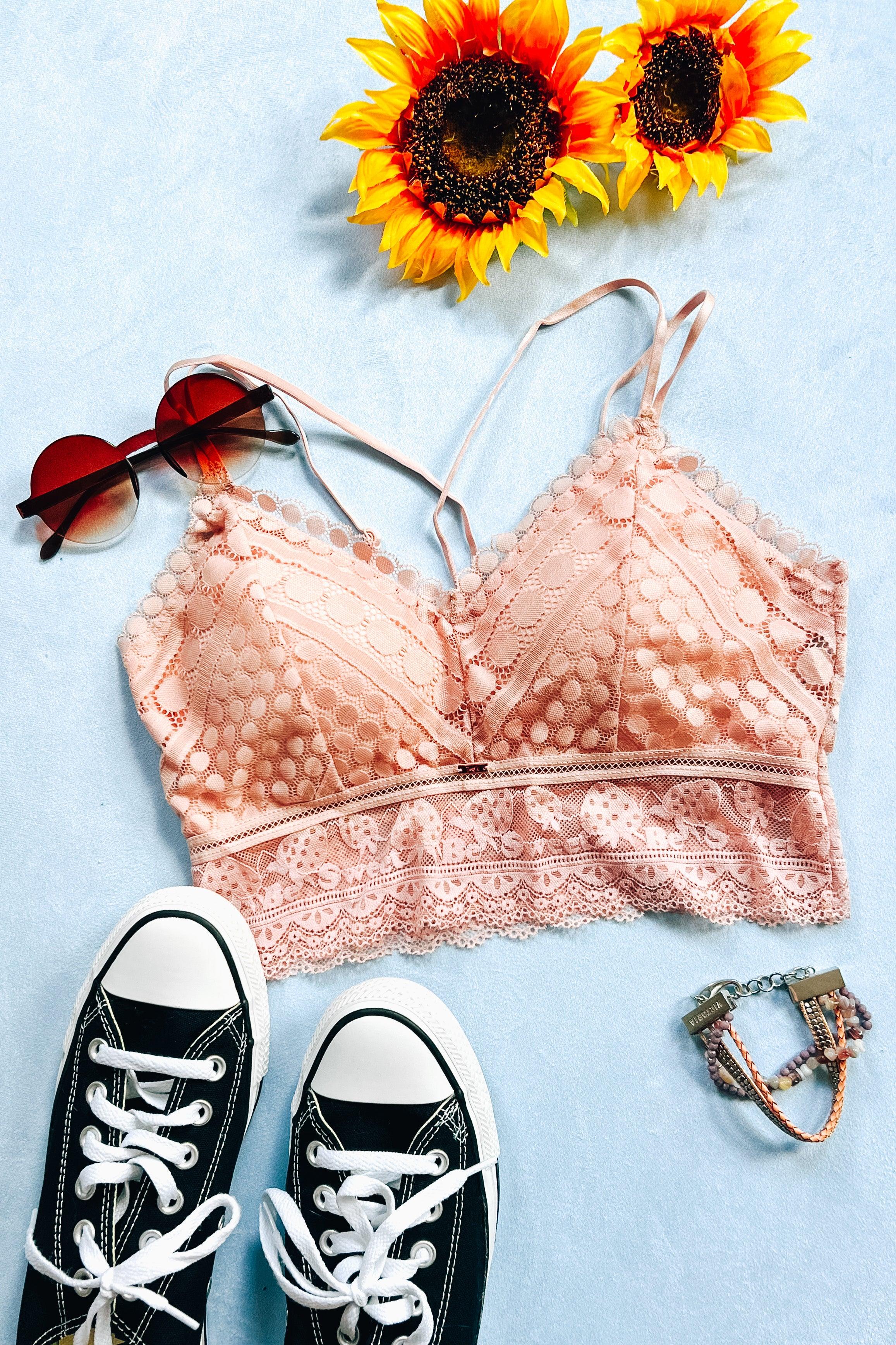 Light blush lace bralette with intricate circular and floral patterns styled alongside black canvas sneakers, heart-shaped sunglasses, beaded bracelet, and sunflowers on a soft blue background.