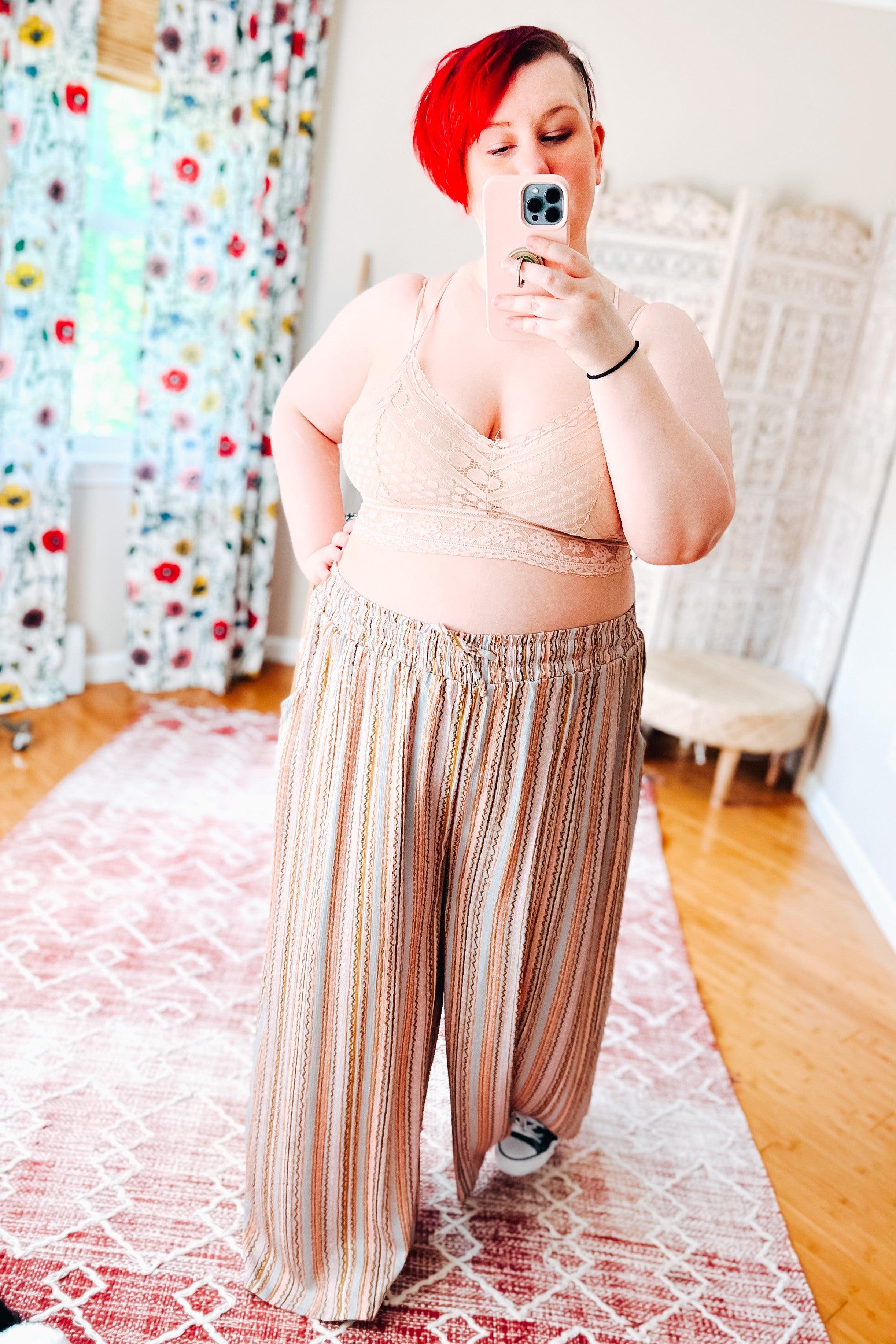Woman with short vibrant red hair wearing a blush lace bralette with delicate patterns paired with high-waisted wide-leg striped pants, taking a mirror selfie in a bright room with floral curtains and wooden floor.