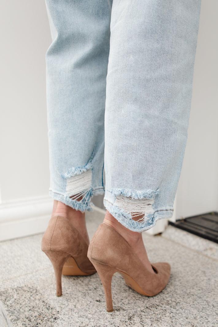 Light blue distressed jeans with frayed ripped hems, paired with taupe suede high-heeled pumps, shown from the back standing indoors on stone steps.