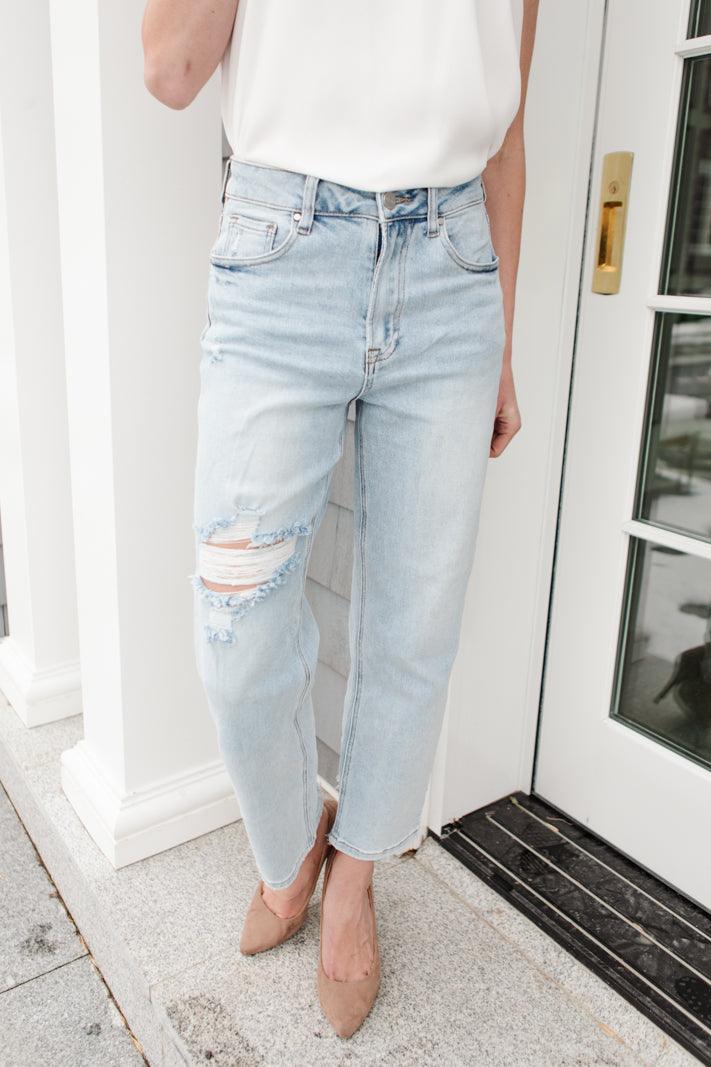 Light blue high-rise jeans with distressed detailing on one thigh, paired with a white sleeveless top and beige pointed heels indoors by a door.