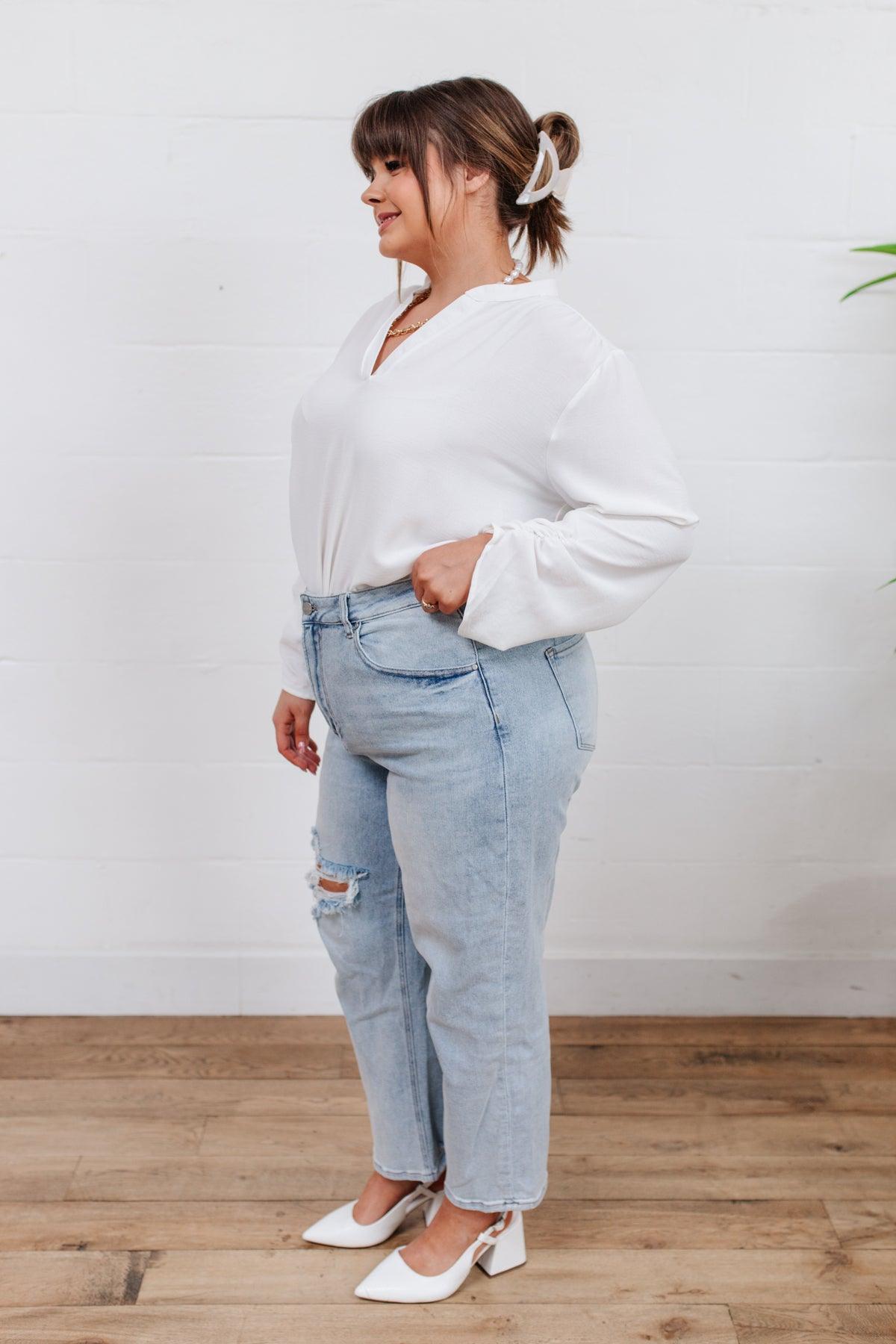 Woman wearing light blue distressed jeans with a rip on one knee paired with a loose white blouse and white low block heels standing indoors