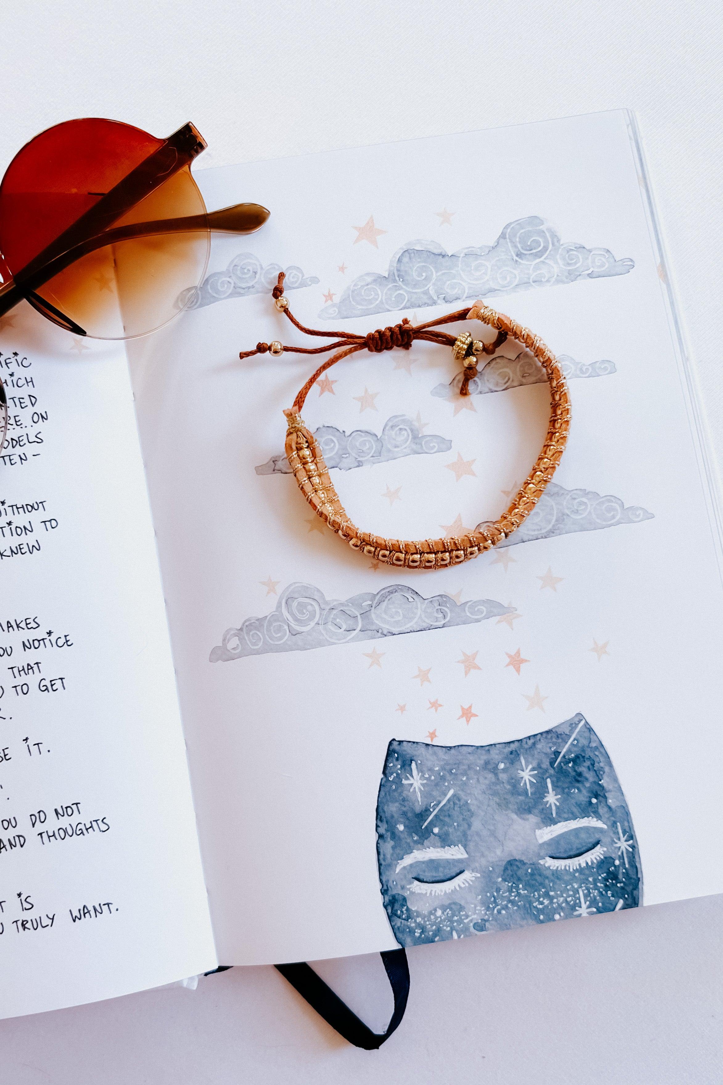 Brown woven bracelet with adjustable beads styled on an open illustrated notebook featuring clouds and stars