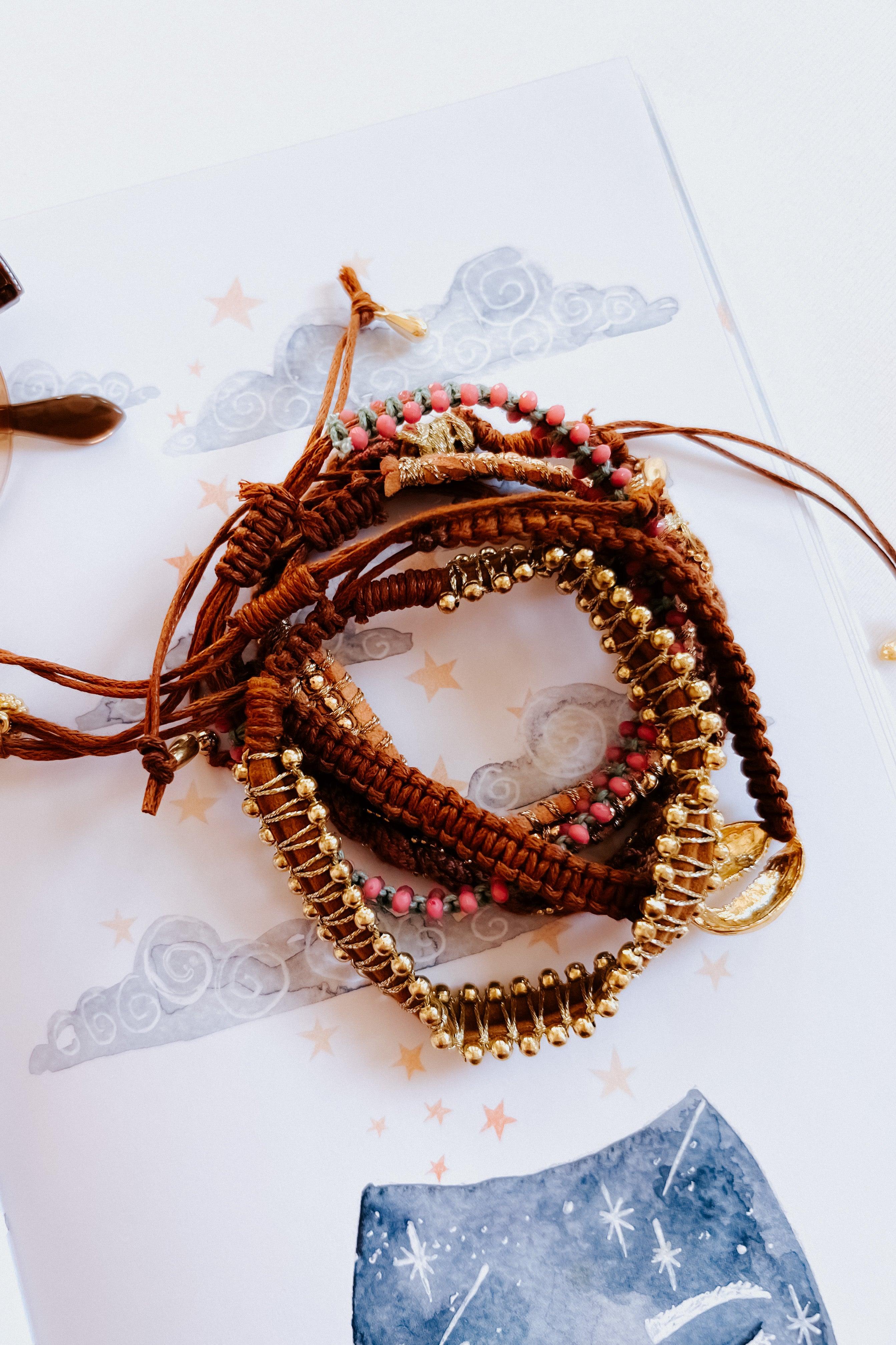 Eclectic brown woven stackable bracelet set with pink beads and gold accents styled on illustrated paper background