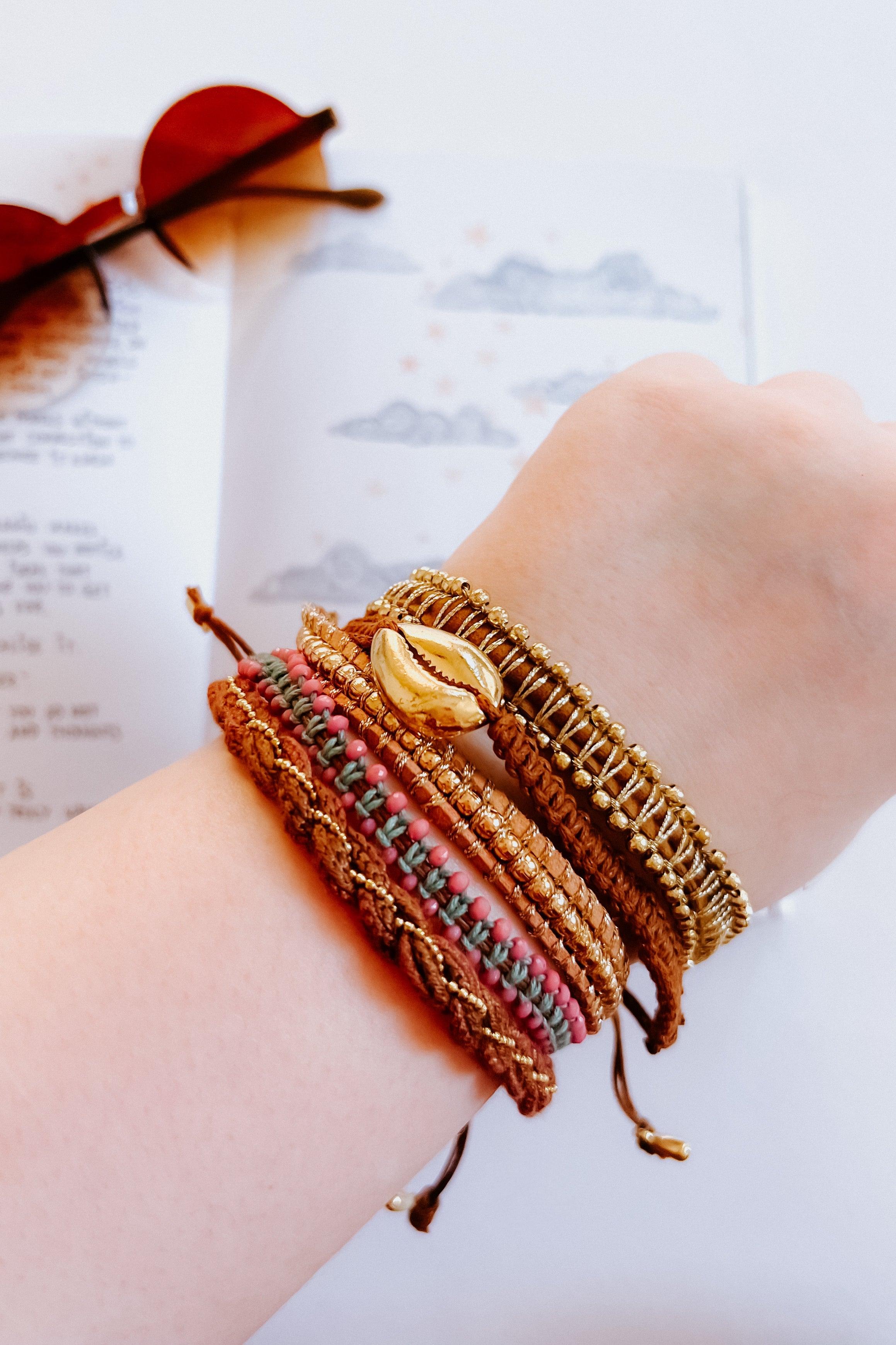 Eclectic stackable brown woven bracelets with beads and gold shell charm on wrist over illustrated clouds and stars background