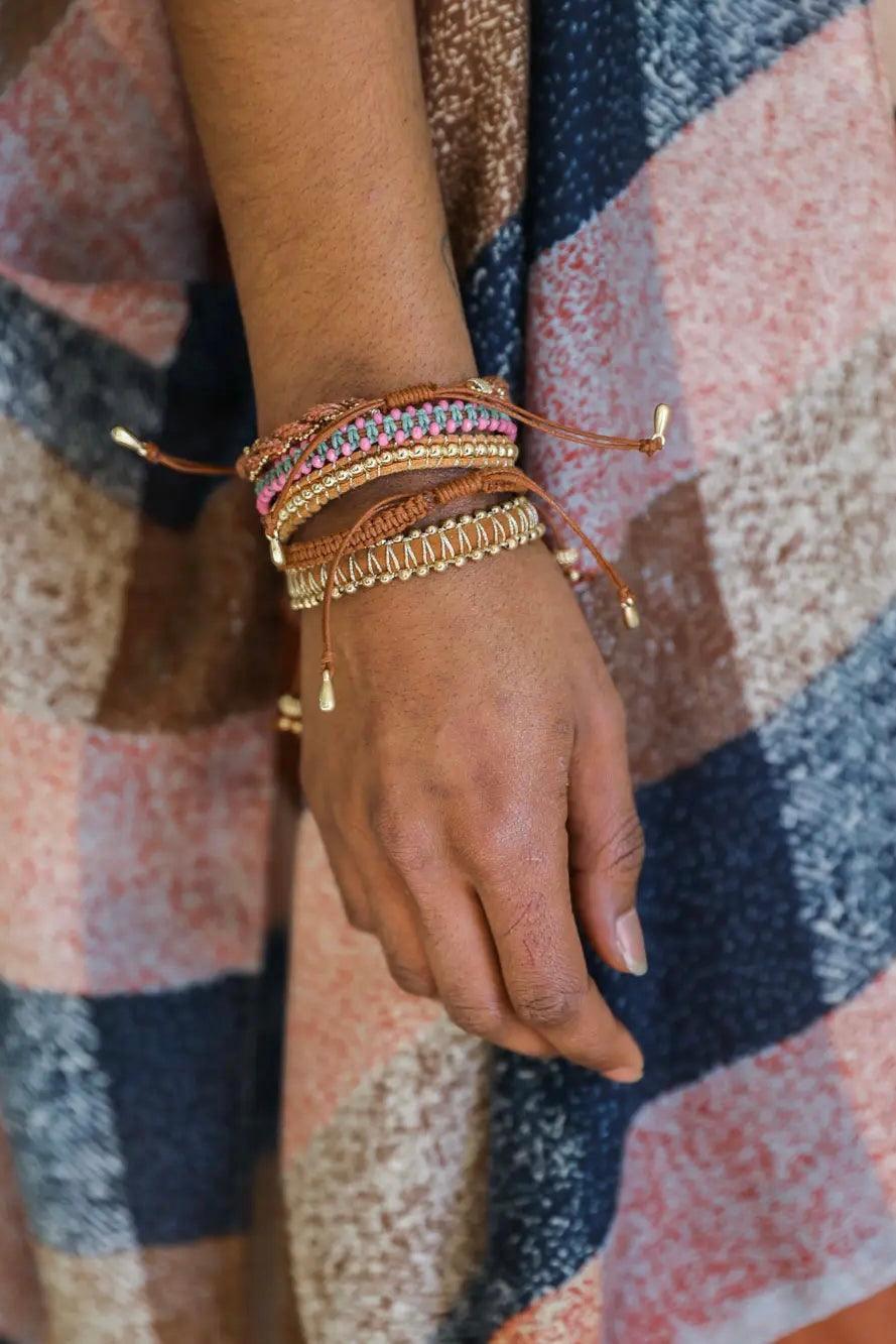 Eclectic brown woven and beaded bracelets with pink and gold accents styled on wrist over patterned fabric background