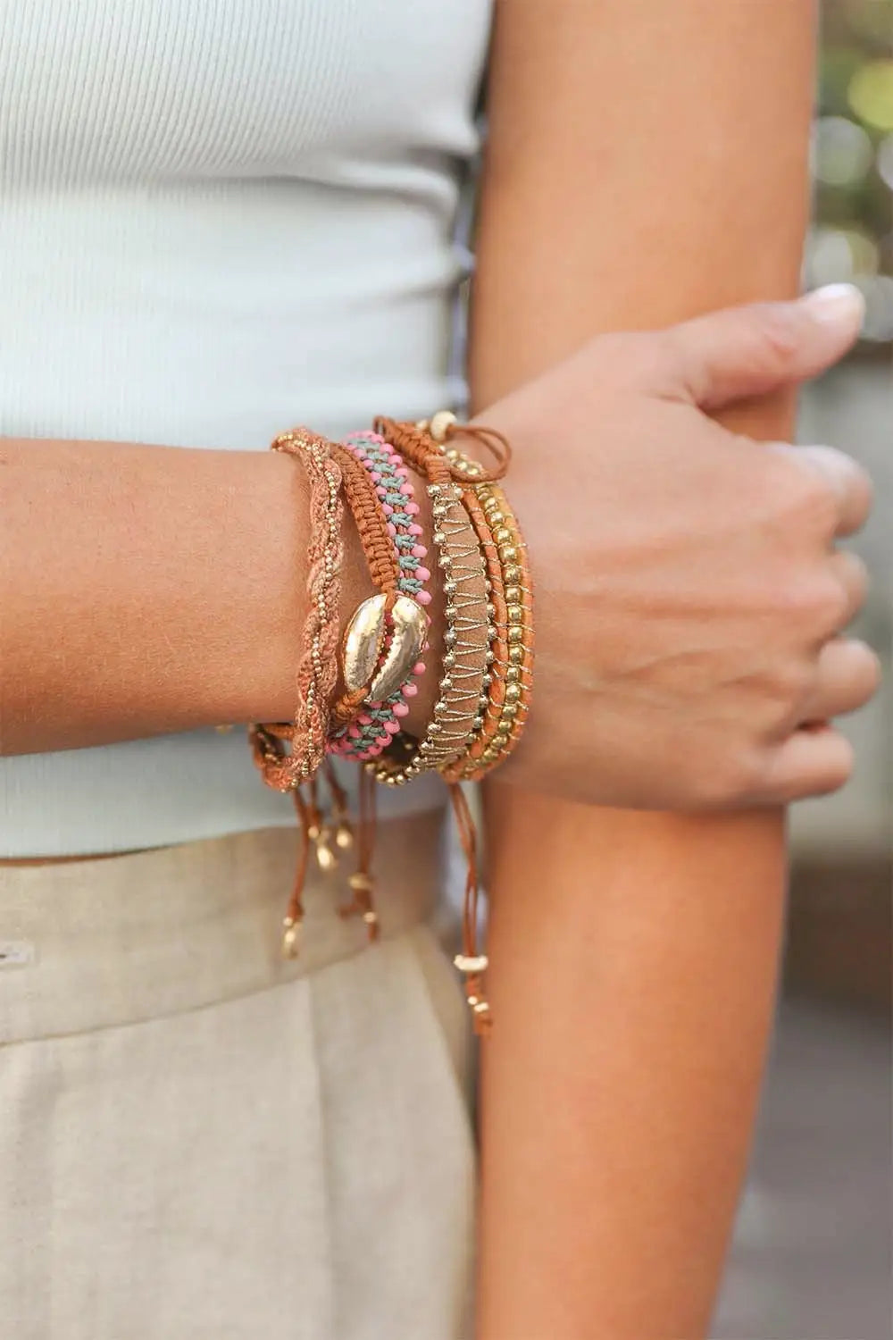 Brown woven stackable beaded bracelets featuring gold accents and a shell charm styled on a wrist with neutral clothing