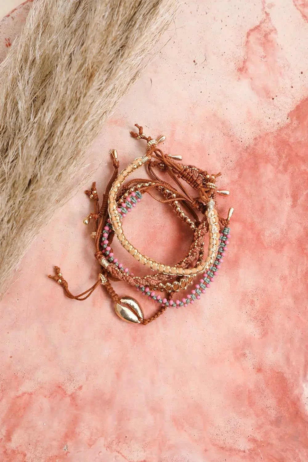 Brown woven stackable bracelets with beads and gold accents styled on a pink marbled surface with dried pampas grass
