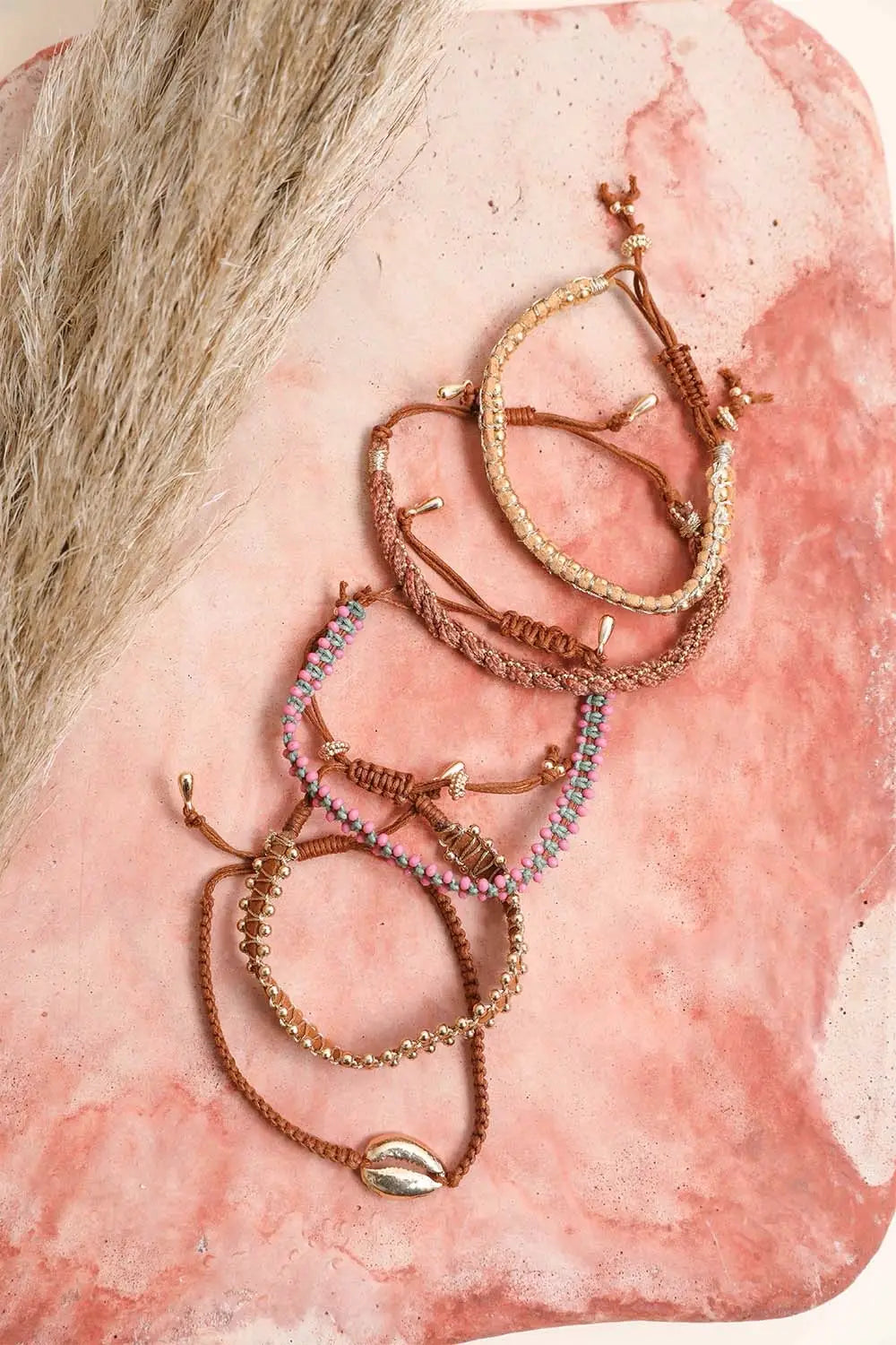 Eclectic brown beaded and woven stackable bracelets with gold accents styled on a pink marbled surface with pampas grass