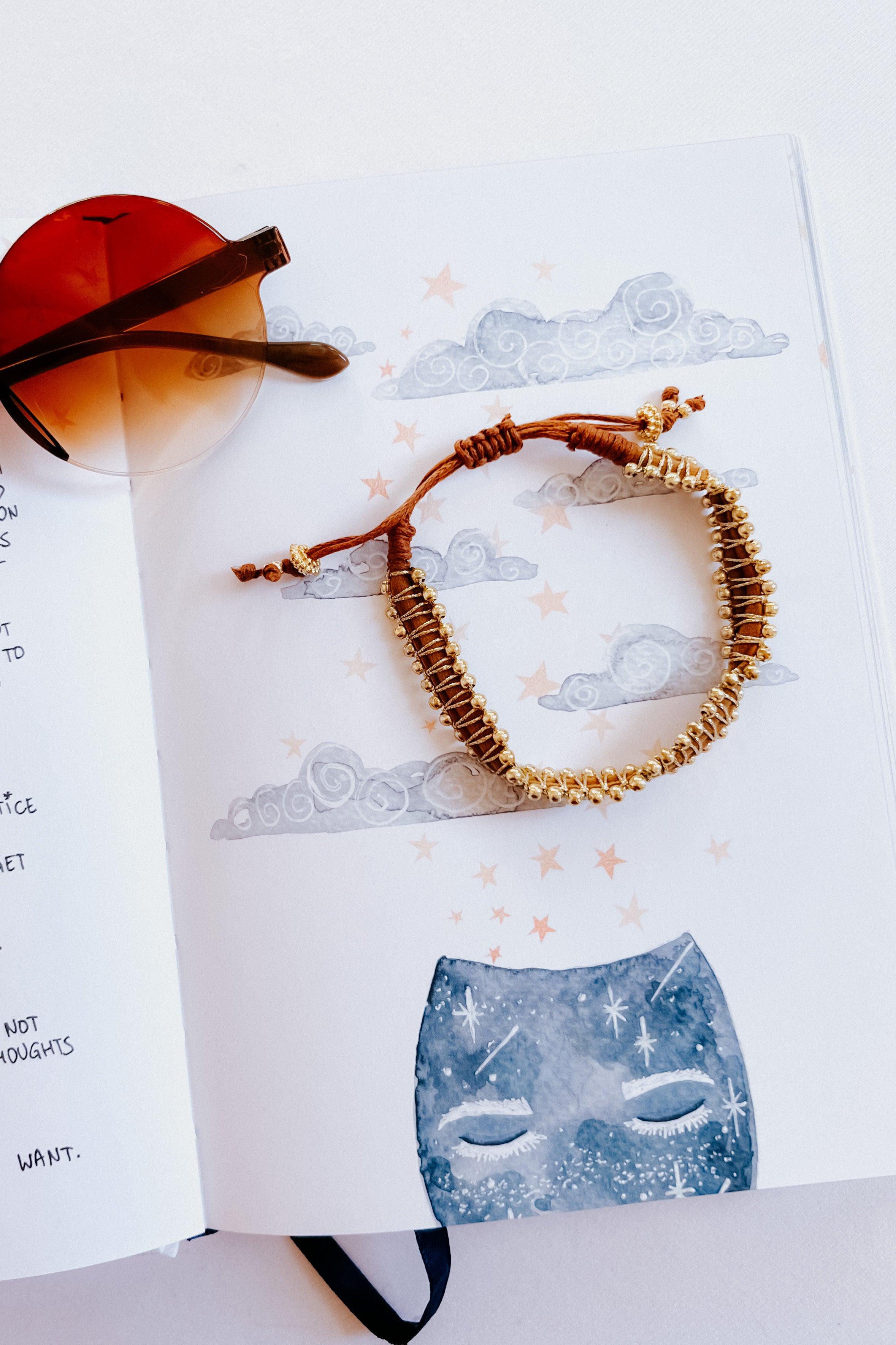 Brown woven beaded bracelet with gold accents styled on an illustrated page with clouds and stars beside amber sunglasses