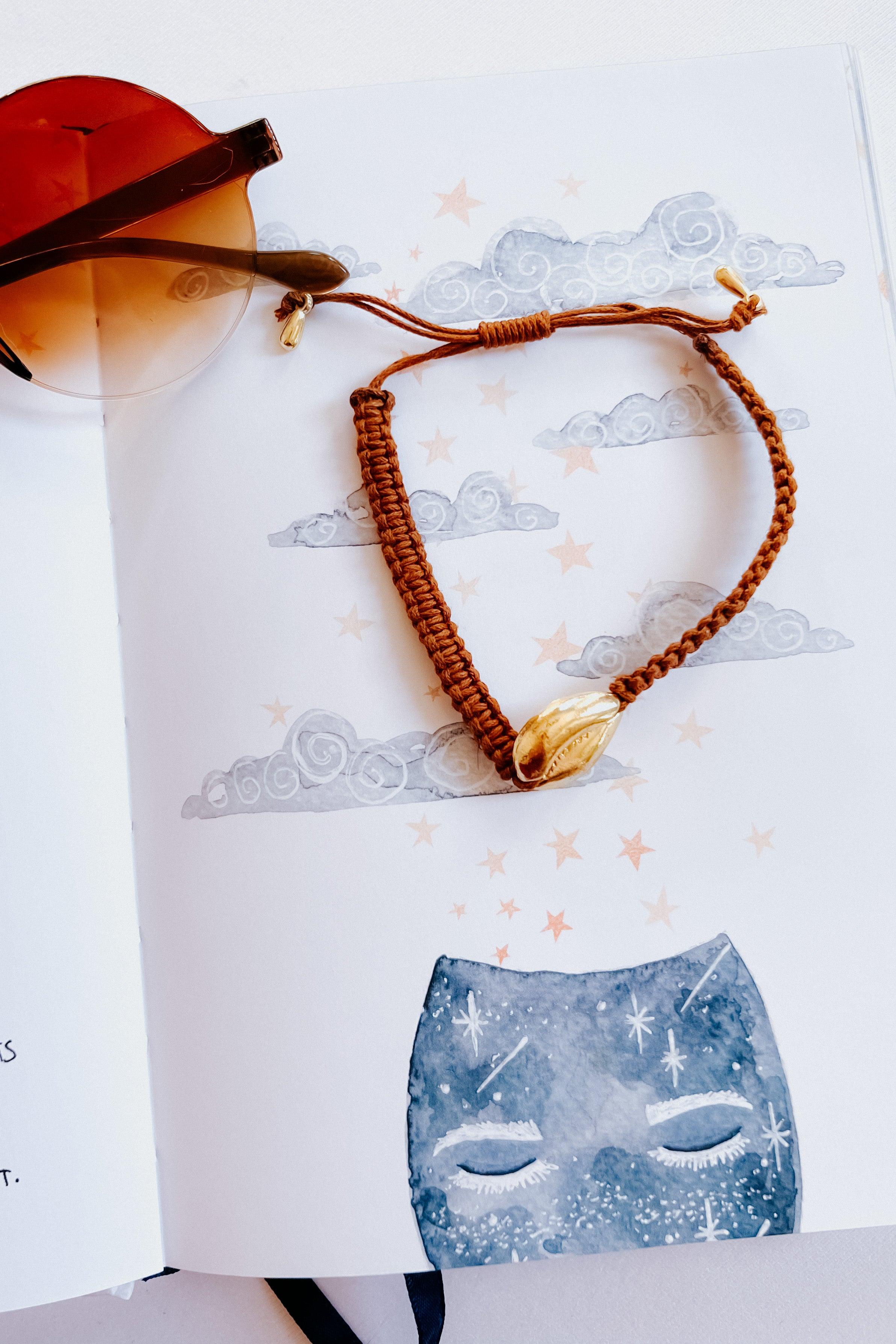 Brown woven bracelet with gold shell charm styled on a whimsical illustrated page with clouds and stars nearby