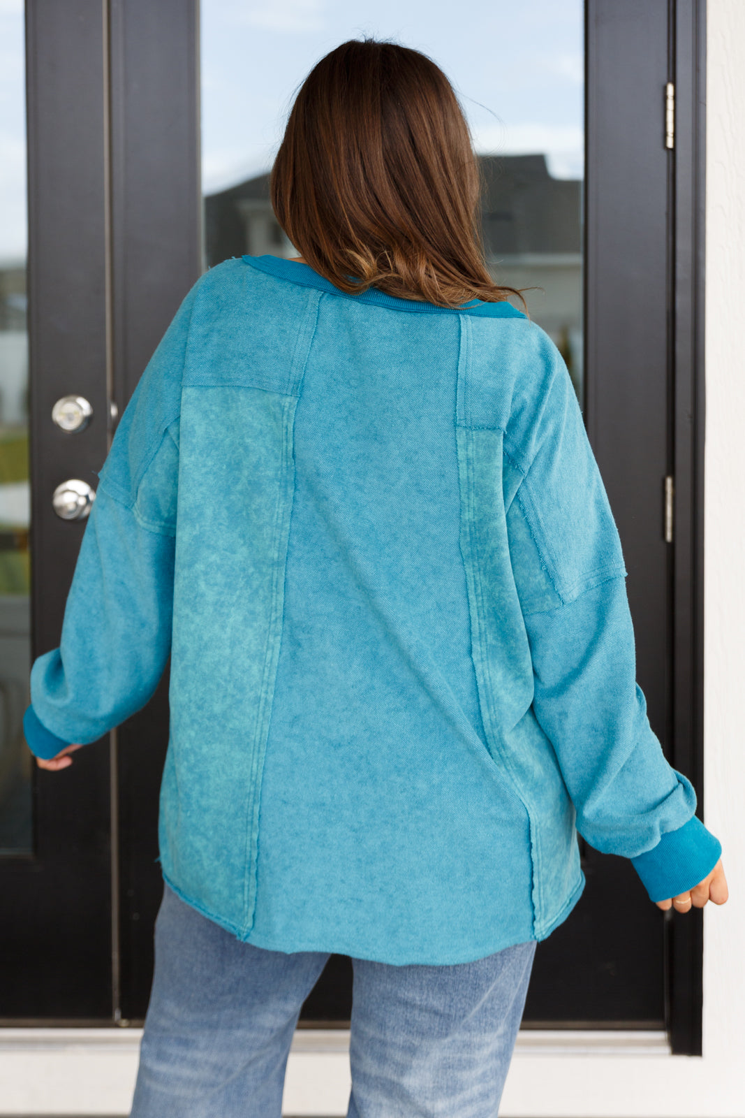A woman with her back facing the camera wearing a blue mineral wash sweater over blue jeans against an outdoor setting.