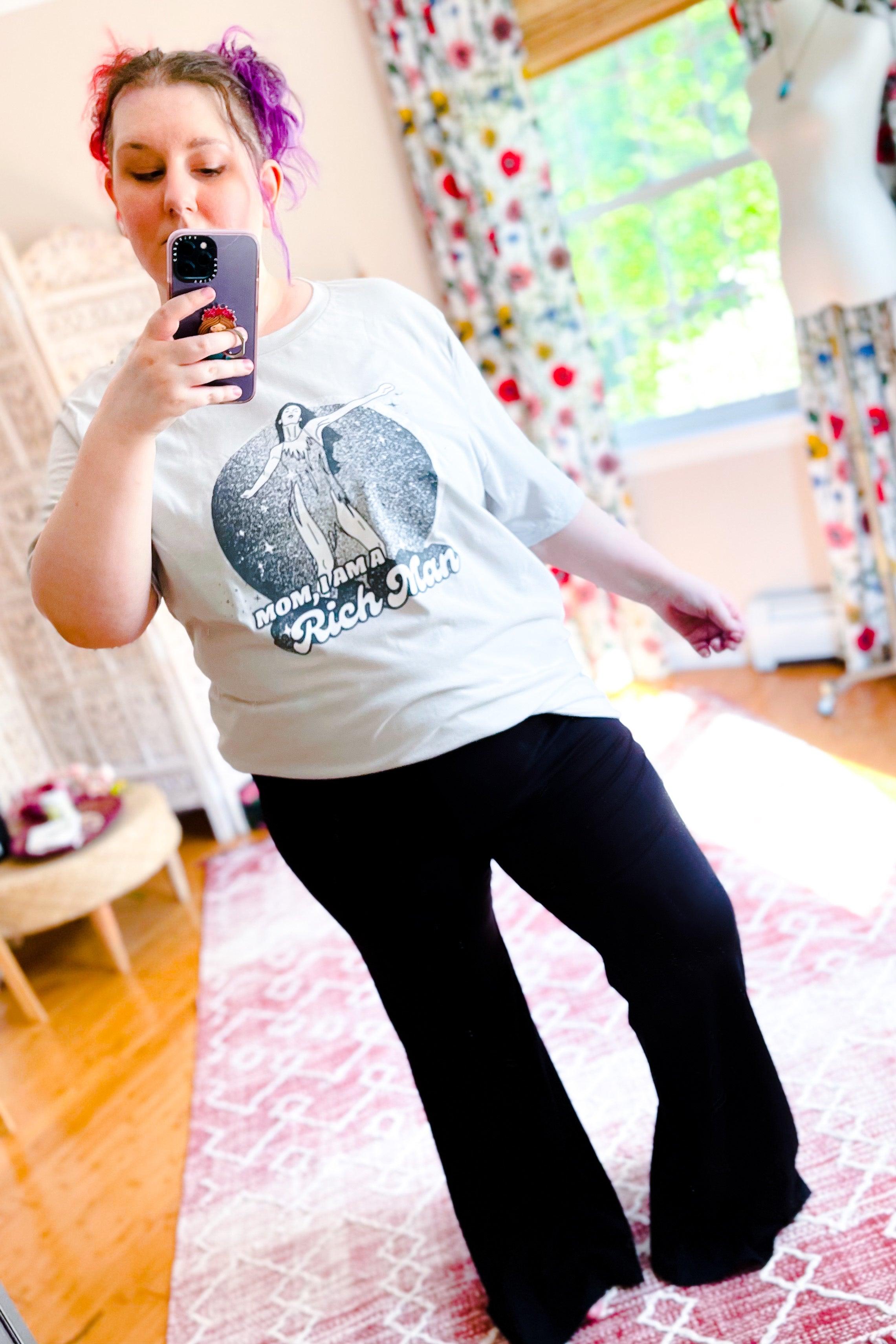 Woman with purple hair wearing black flared pants and graphic tee taking a mirror selfie in bright cozy room