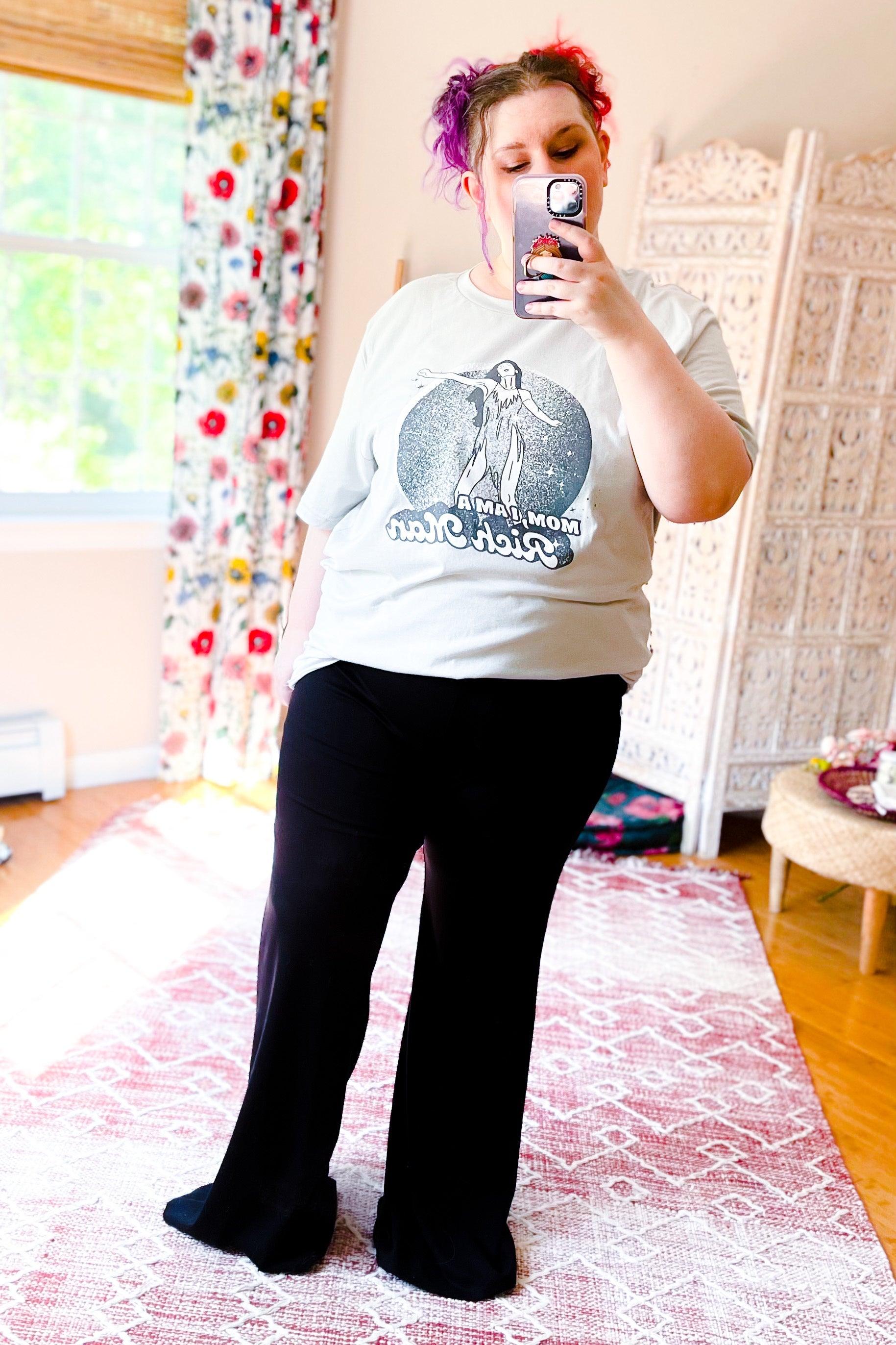 Woman wearing black flared pants with graphic tee and vibrant pink and purple hair taking mirror selfie in bright room