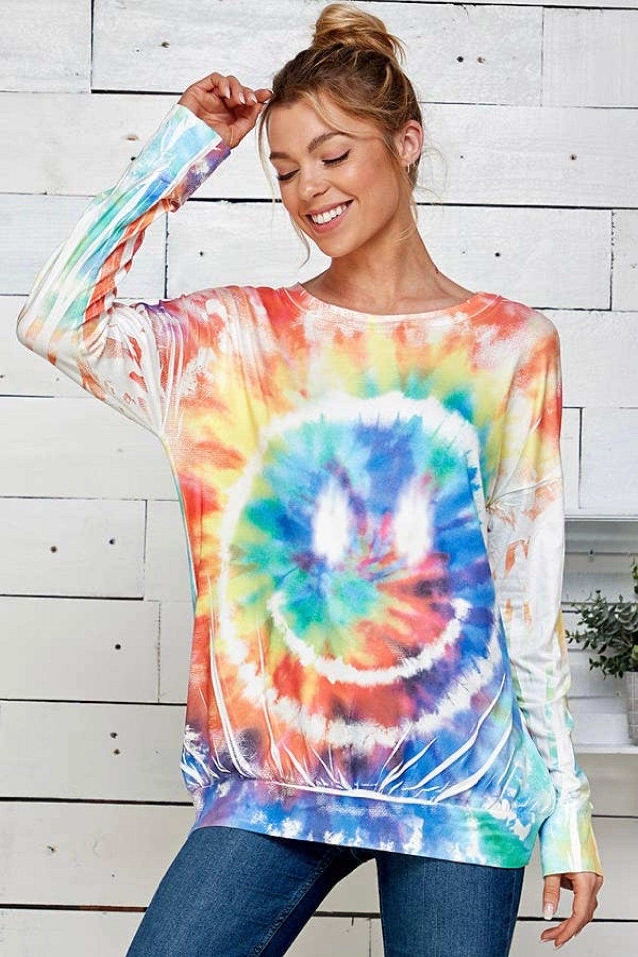 Woman wearing a tie dye rainbow pullover with a cheerful smiley design in a relaxed fit, styled with jeans indoors