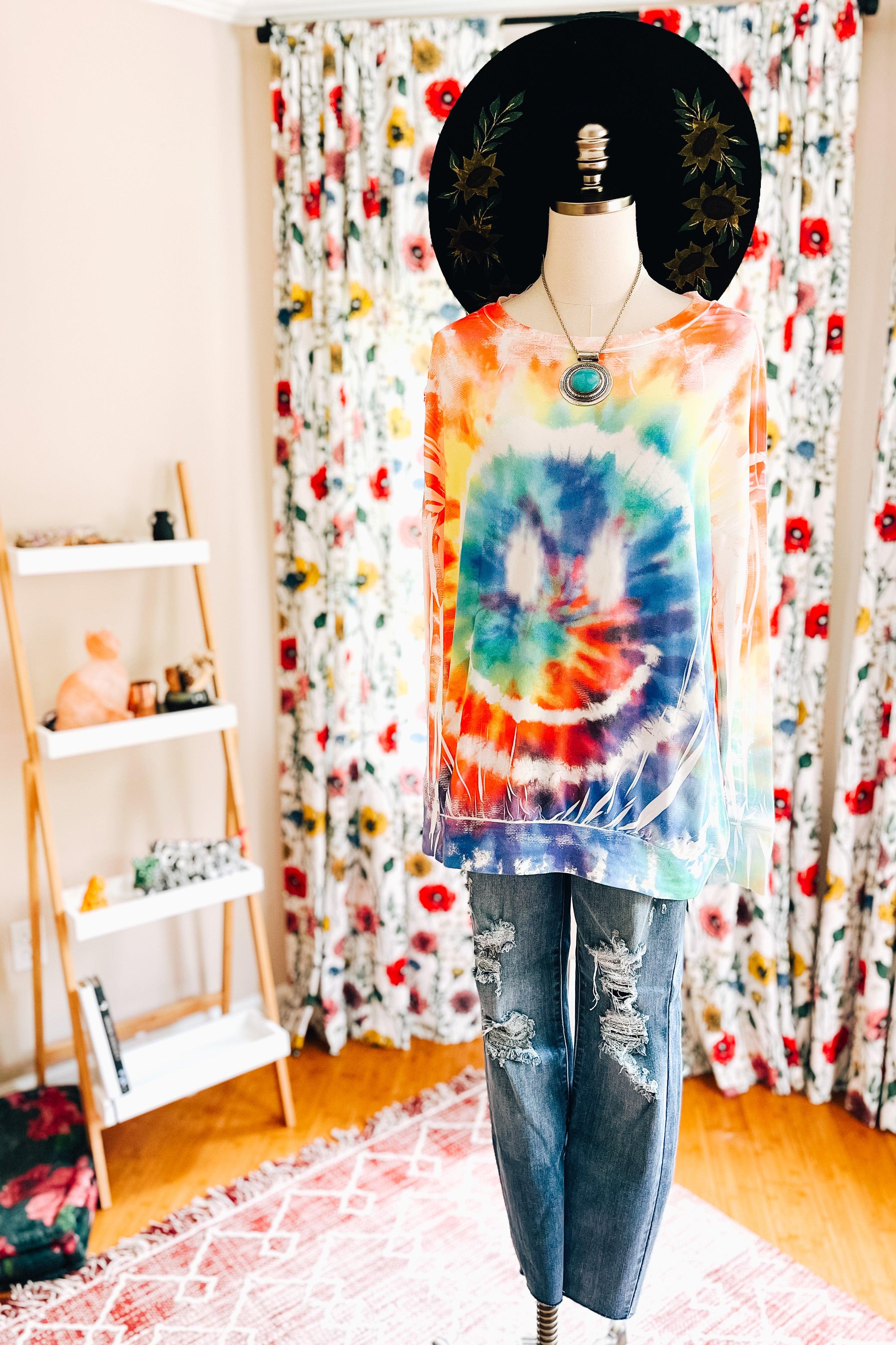 Tie dye rainbow pullover with a large smiley design styled with distressed jeans and a wide-brimmed black hat indoors
