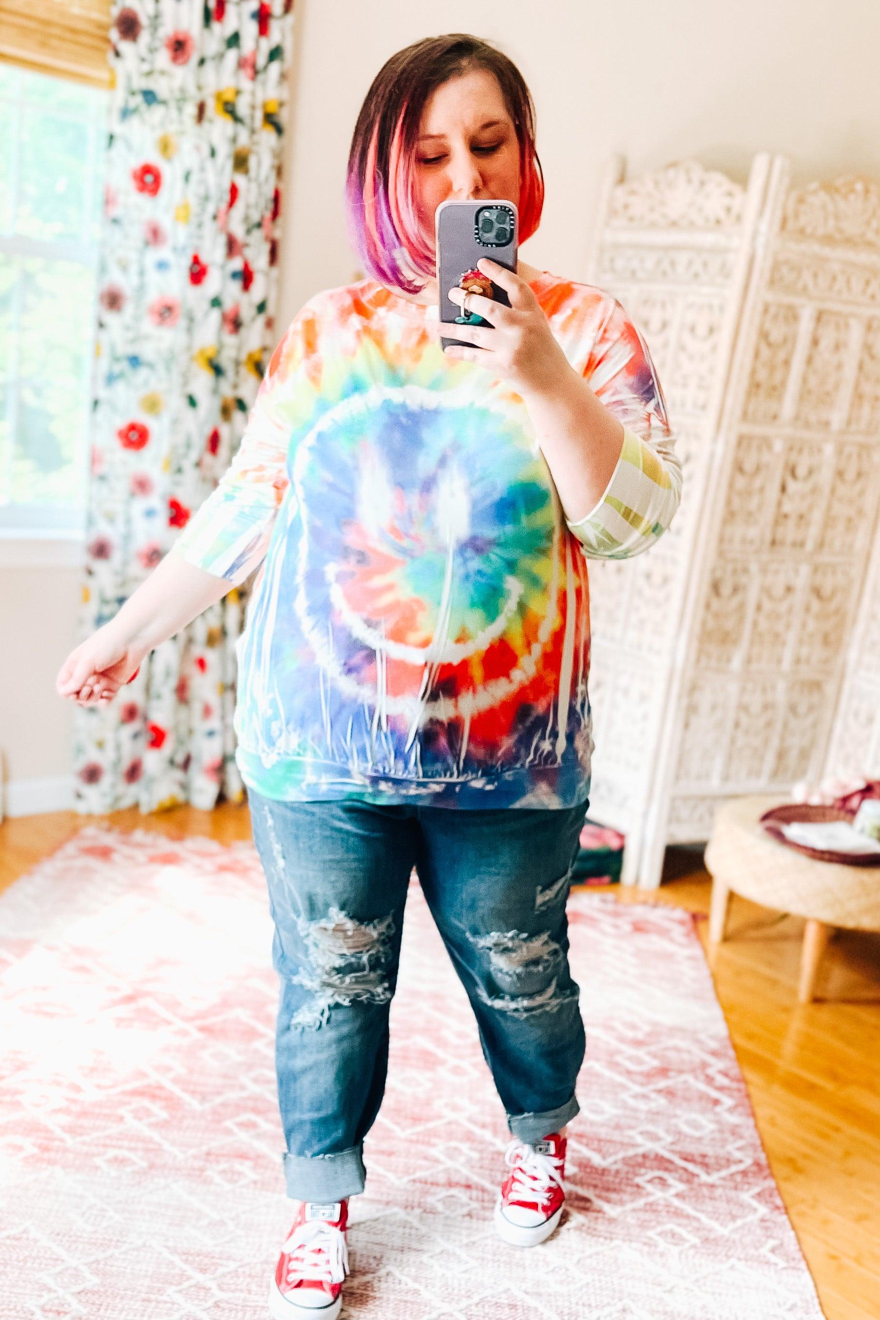 Woman wearing a tie dye rainbow pullover with a bold circular pattern, paired with distressed jeans and red sneakers indoors