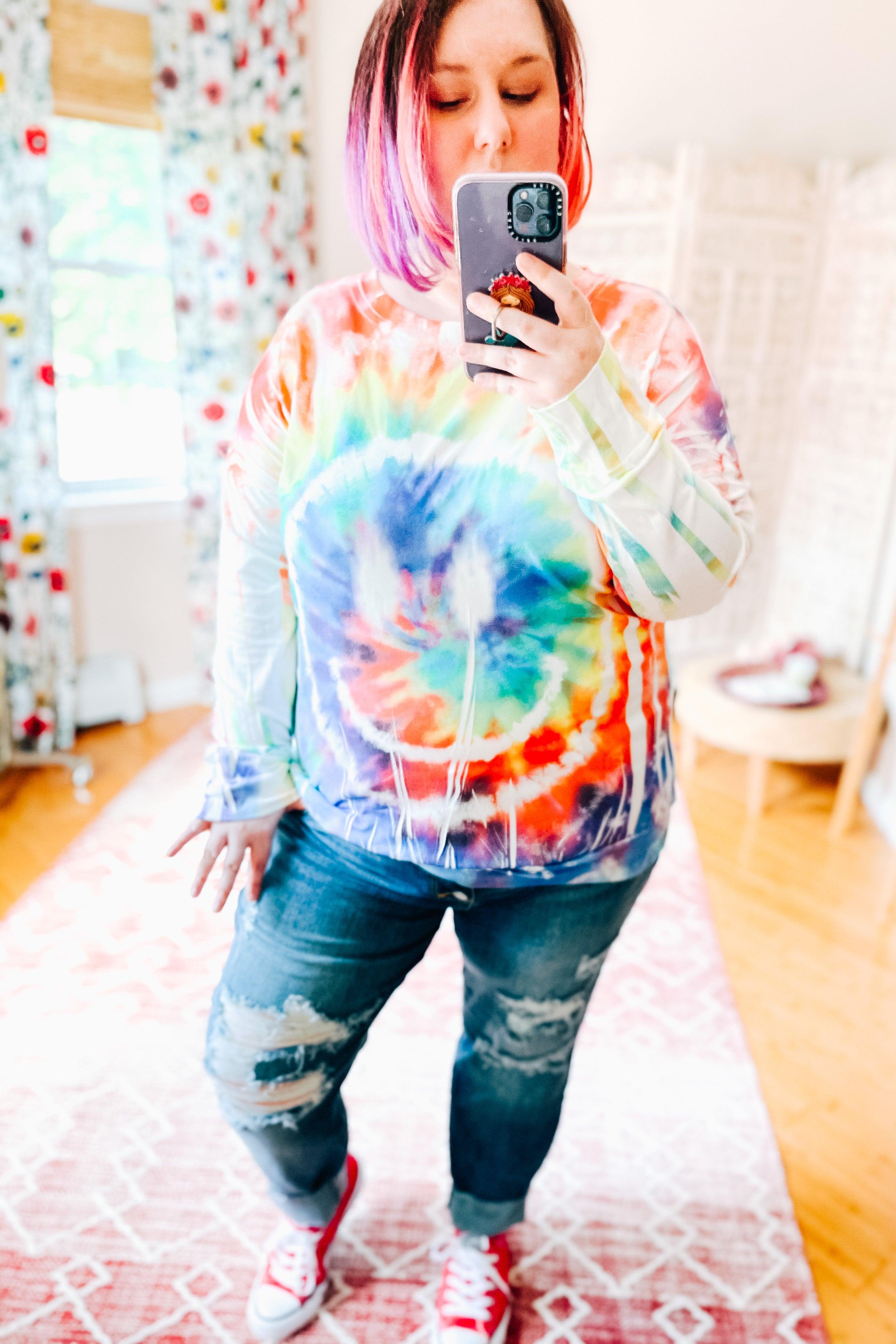 Woman wearing a colorful tie dye rainbow pullover with a bold spiral design paired with ripped jeans and red shoes indoors