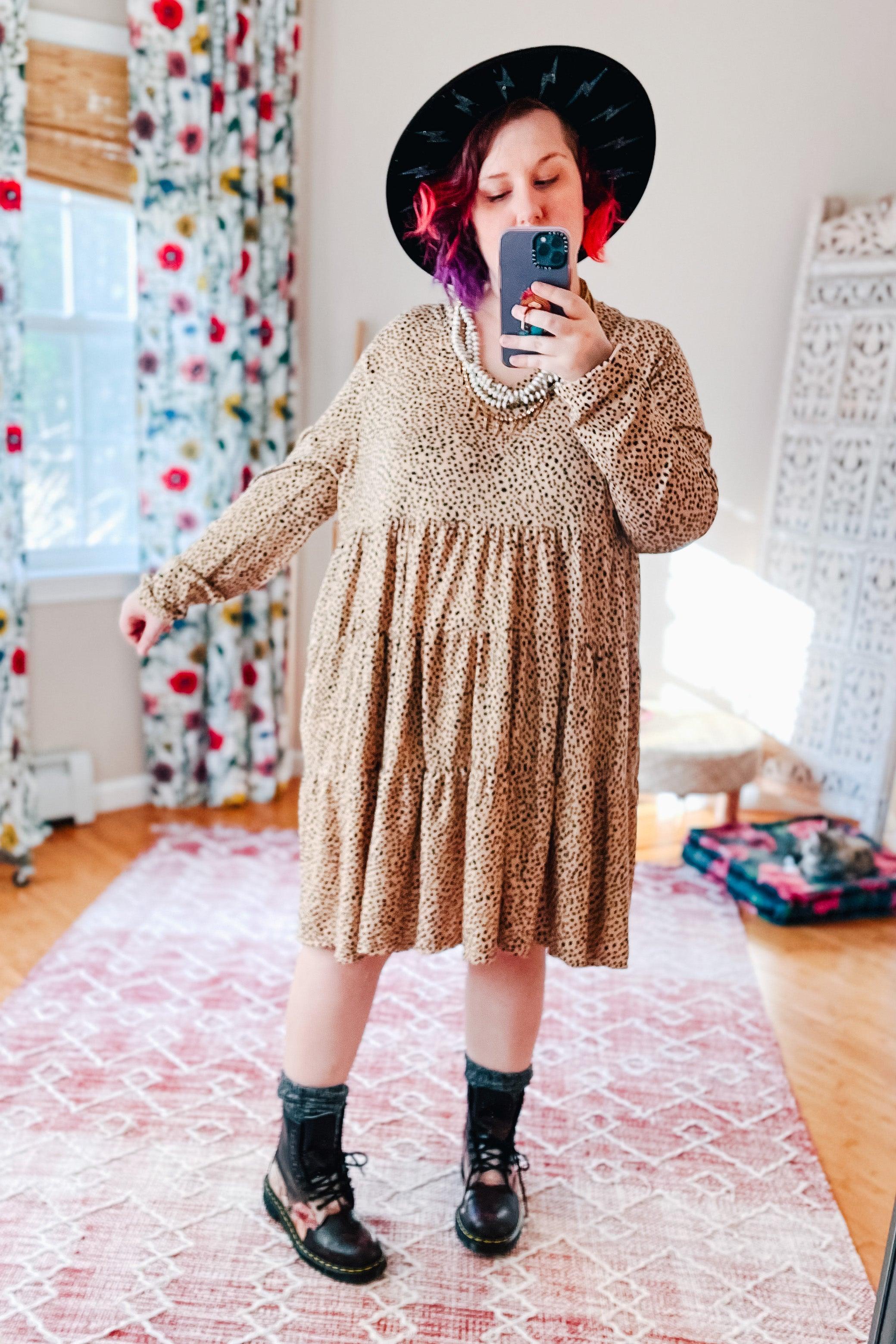 Plus size woman wearing taupe animal print dress with tiered skirt, black patterned boots, black hat, and beaded necklace indoors