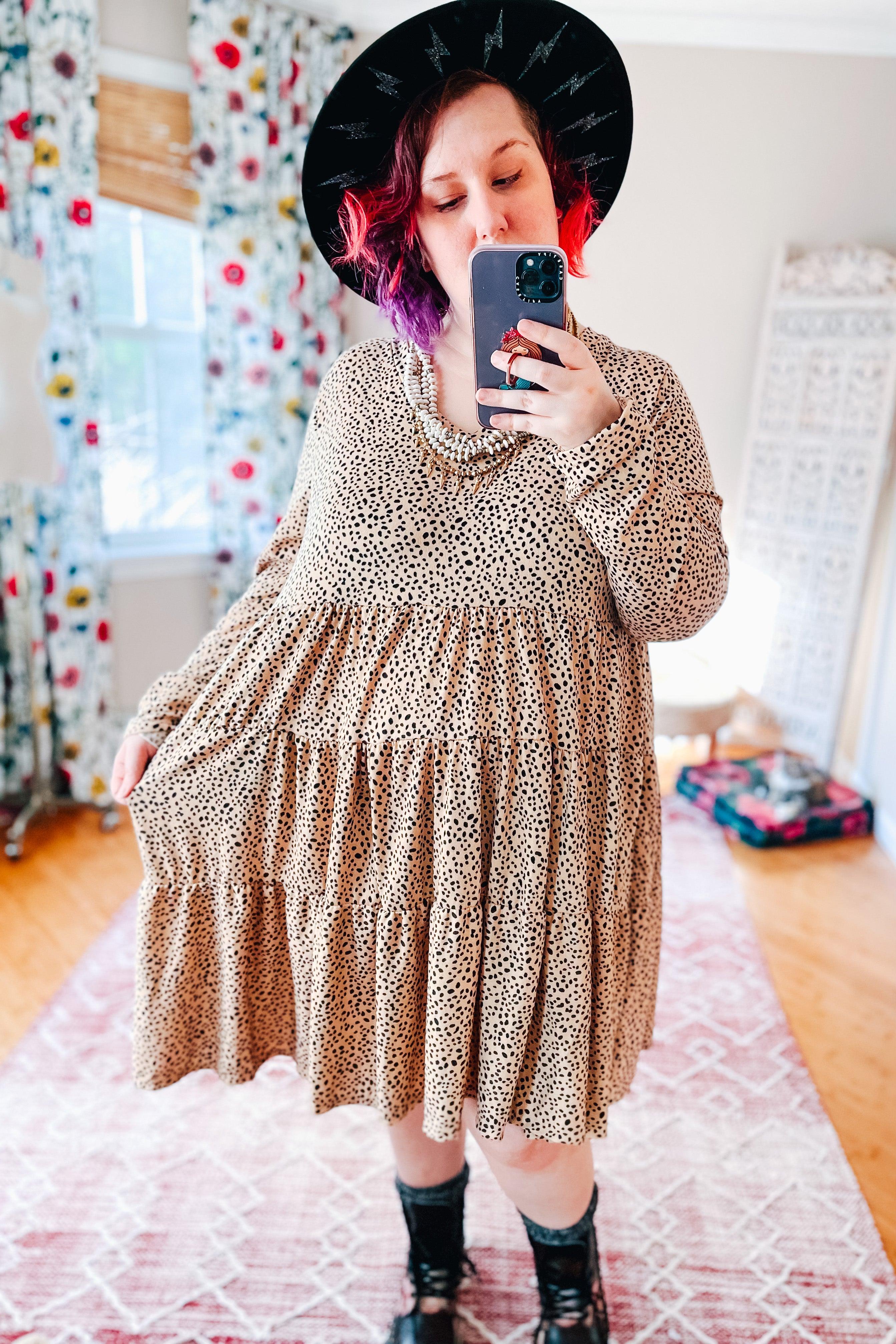 Plus size woman wearing taupe spotted V neck dress with tiered skirt, black boots, wide black hat, and beaded necklace indoors