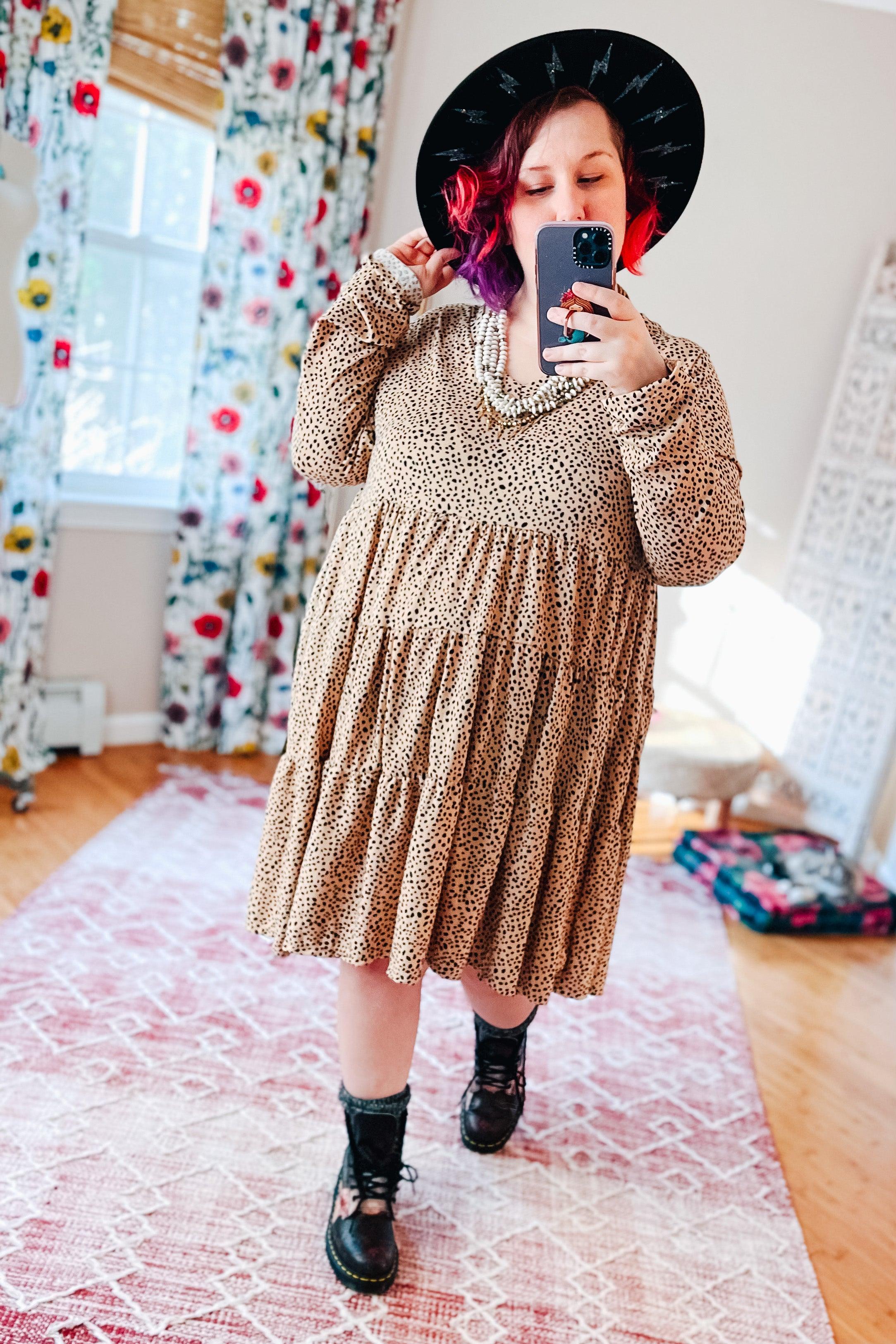 Woman with purple and red hair wearing taupe spotted V neck dress with tiered skirt, black boots, black wide hat, and statement necklace indoors