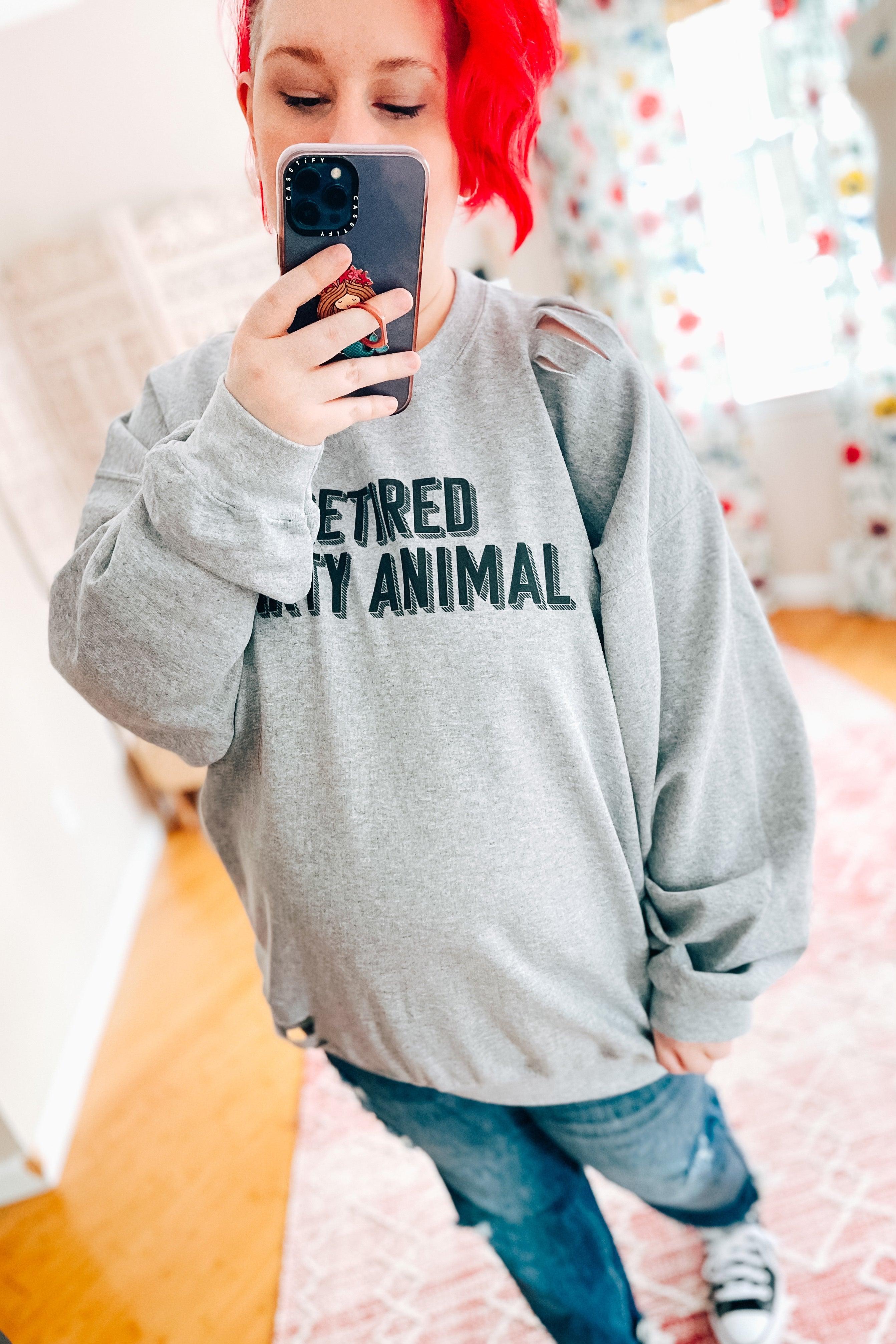 Woman with red hair wearing grey distressed pullover with bold "Retired Party Animal" text taking mirror selfie indoors