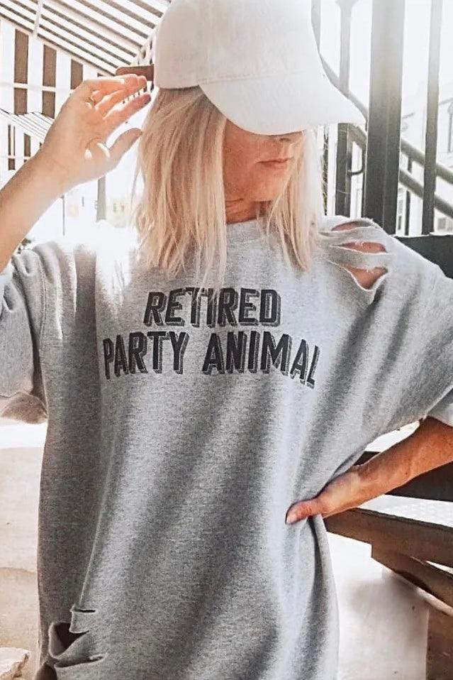 Woman wearing grey distressed pullover with "Retired Party Animal" text and white cap in bright indoor setting