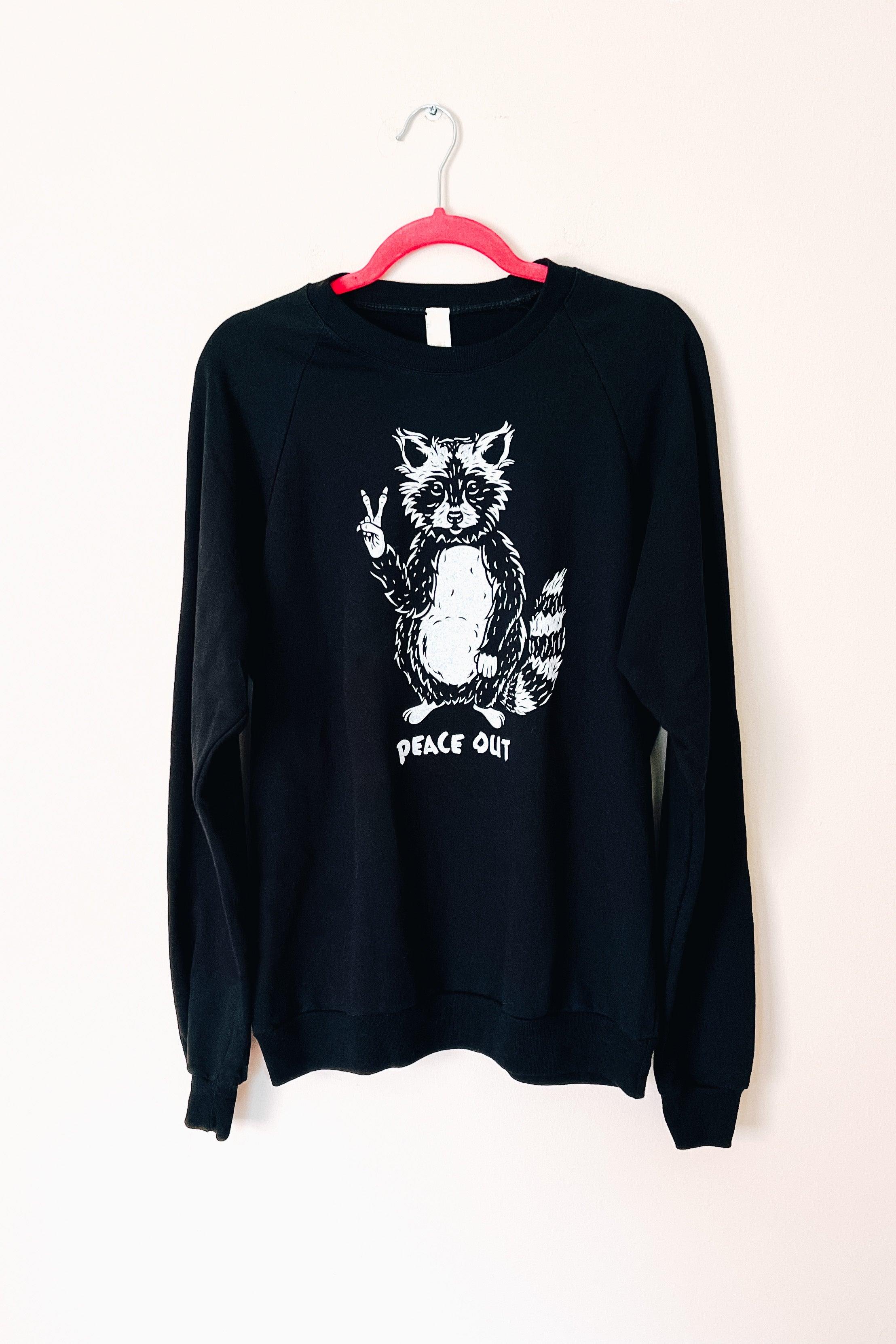 Black organic cotton sweatshirt with raccoon graphic flashing peace sign and “peace out” text beneath design