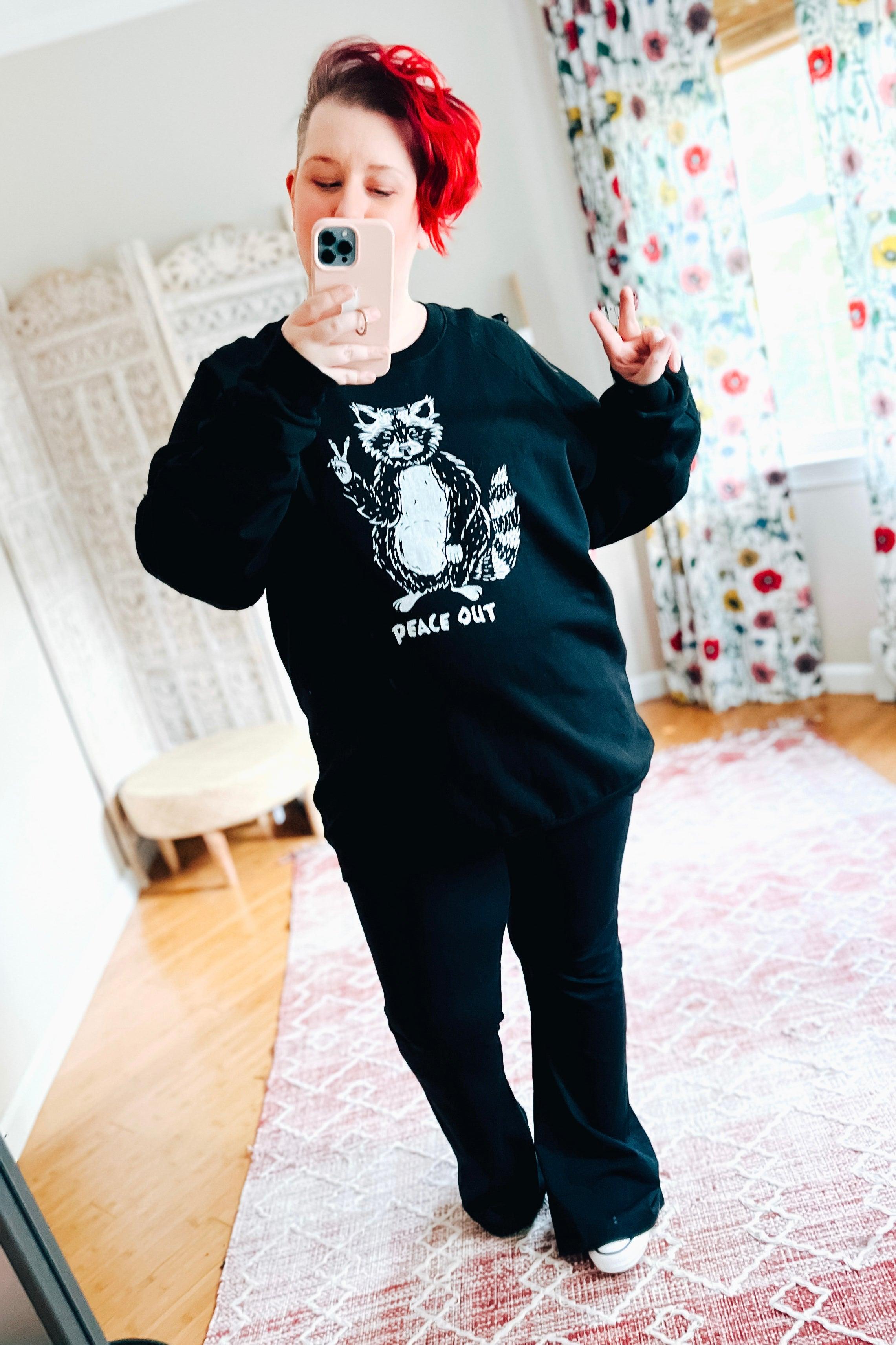 Woman with red hair taking mirror selfie wearing black organic cotton sweatshirt with raccoon peace sign and text "peace out