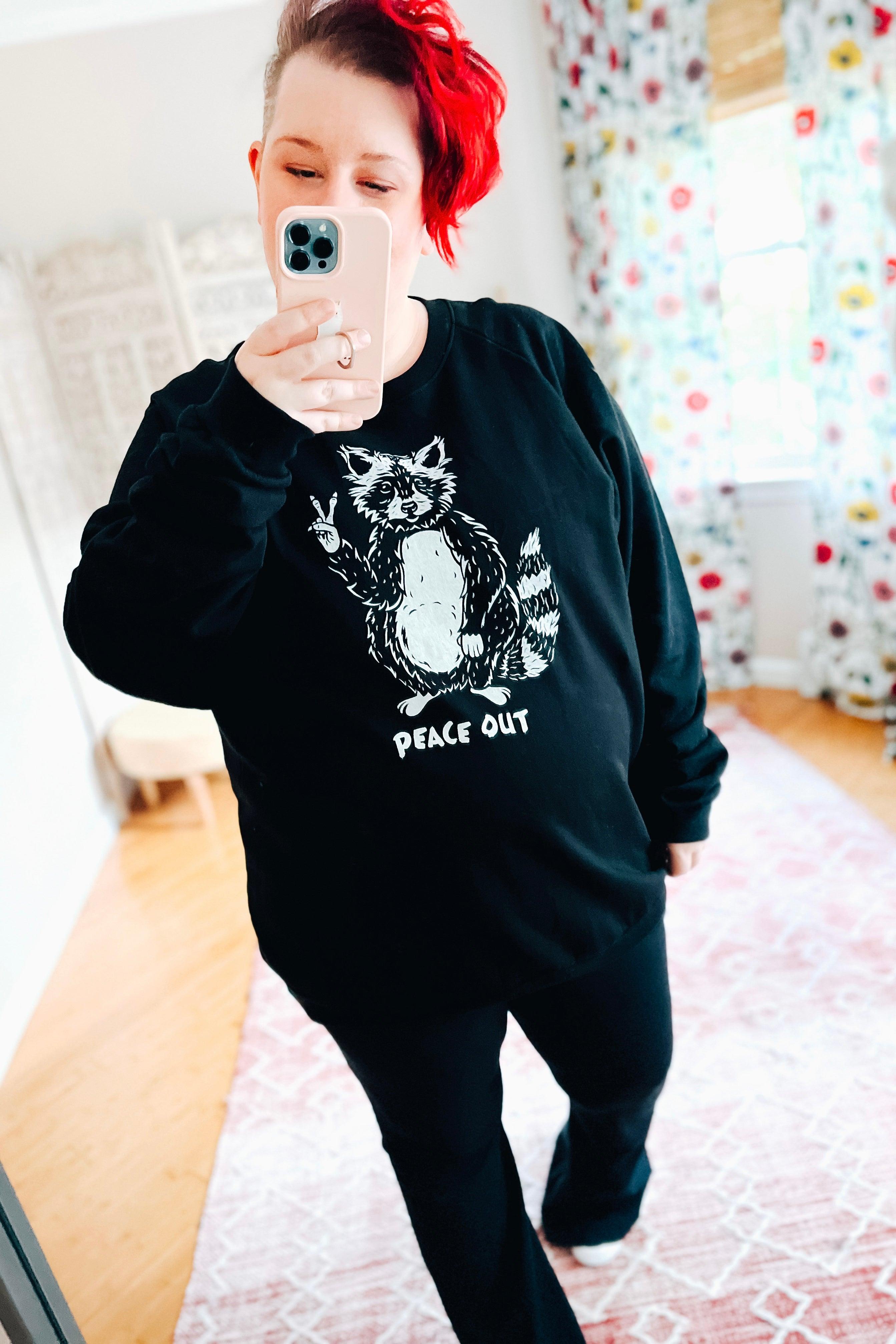 Woman with short red hair taking mirror selfie wearing black organic cotton sweatshirt with raccoon showing peace sign and text below