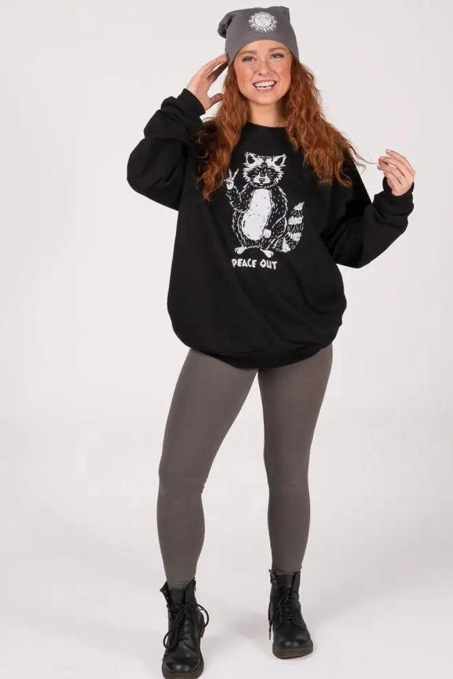 Woman with long red hair wearing black oversized sweatshirt featuring raccoon graphic and "peace out" text paired with gray leggings and boots