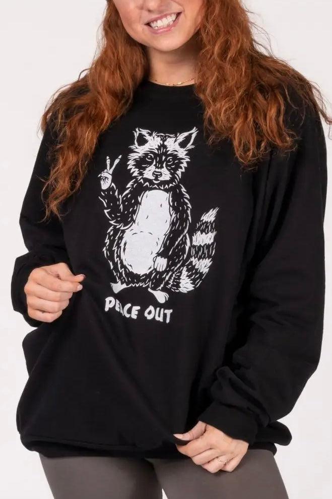 Woman with long red hair wearing black sweatshirt featuring raccoon illustration giving peace sign and text below