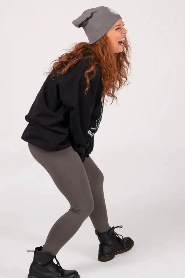 Woman with curly red hair wearing black organic cotton sweatshirt, gray leggings, gray beanie, and black boots laughing side view