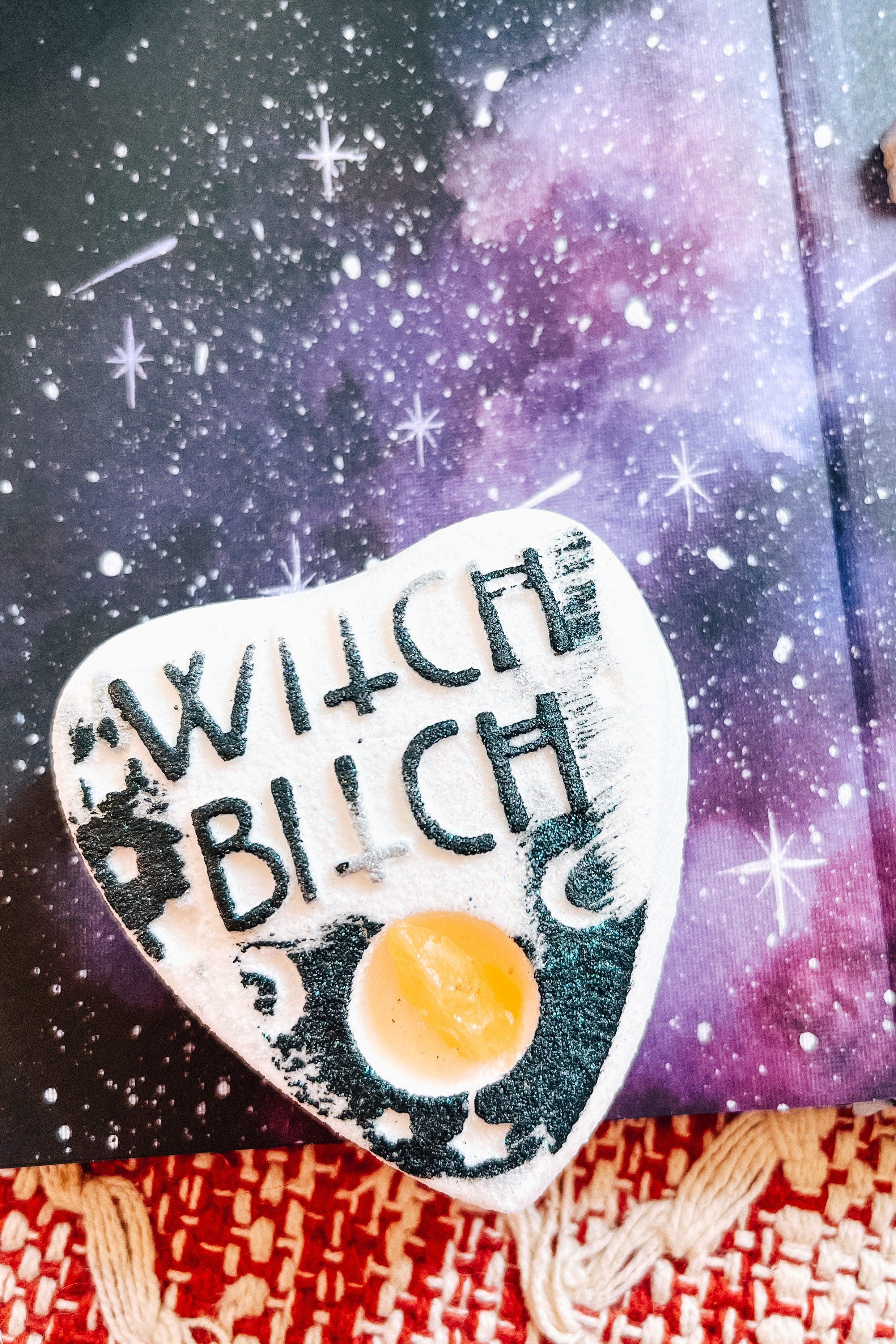 White planchette bath bomb with black lettering “witch bitch” and a citrine crystal set on a purple starry book background and red woven fabric